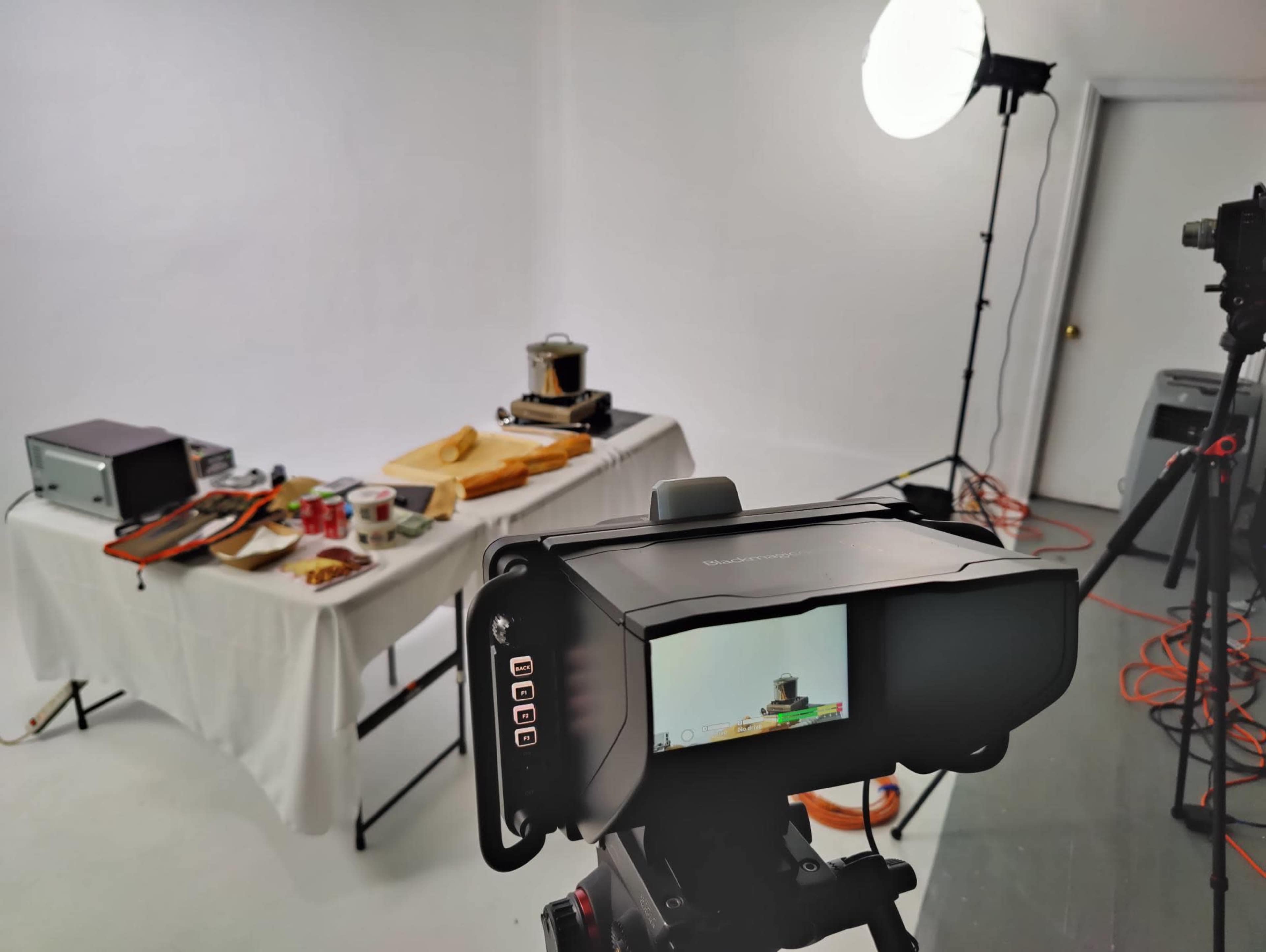 A camera is set up in a studio with a light source, capturing a table displaying various kitchen items and ingredients.