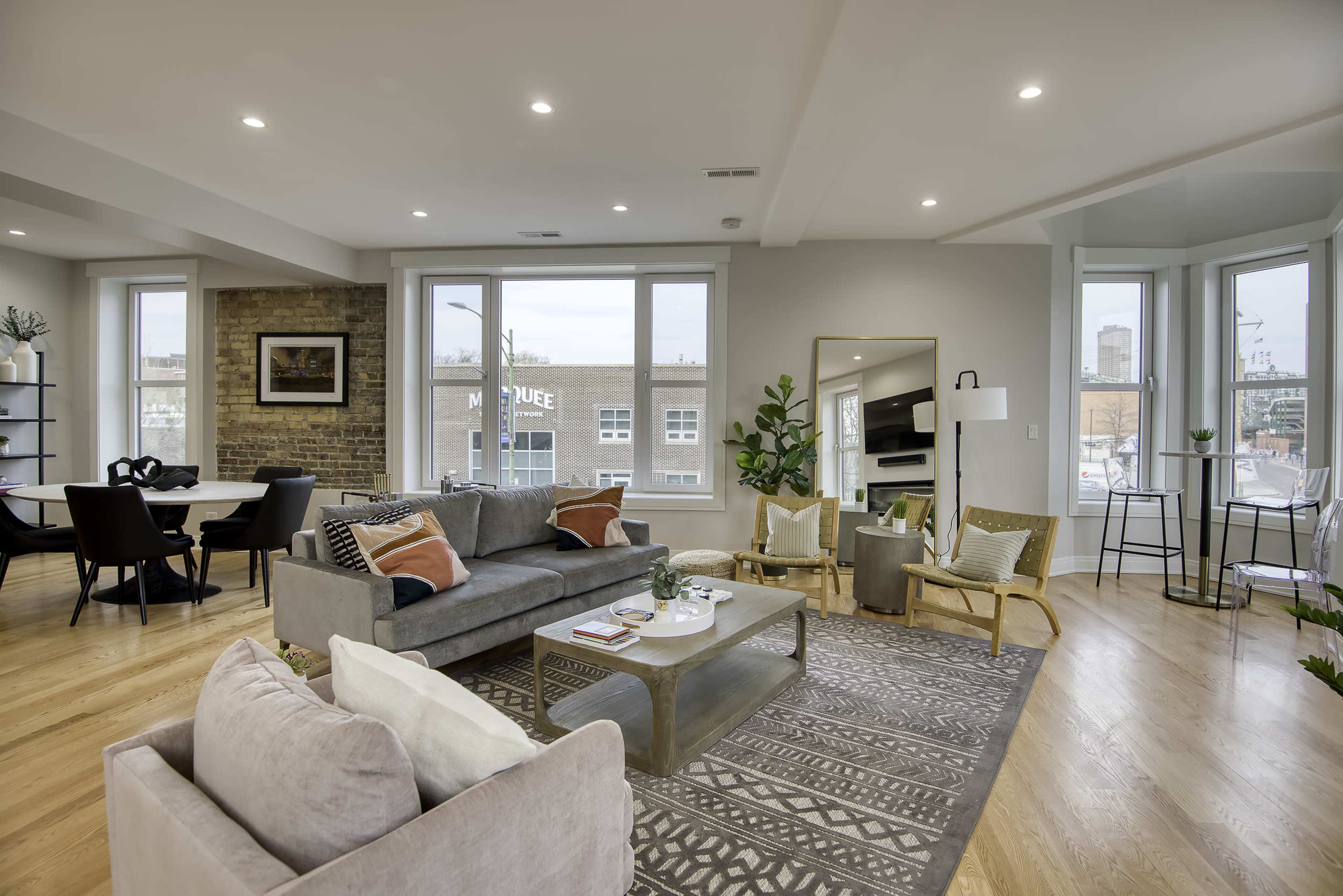 A modern living room with a gray sofa, wooden chairs, and large windows that allow natural light to fill the space.