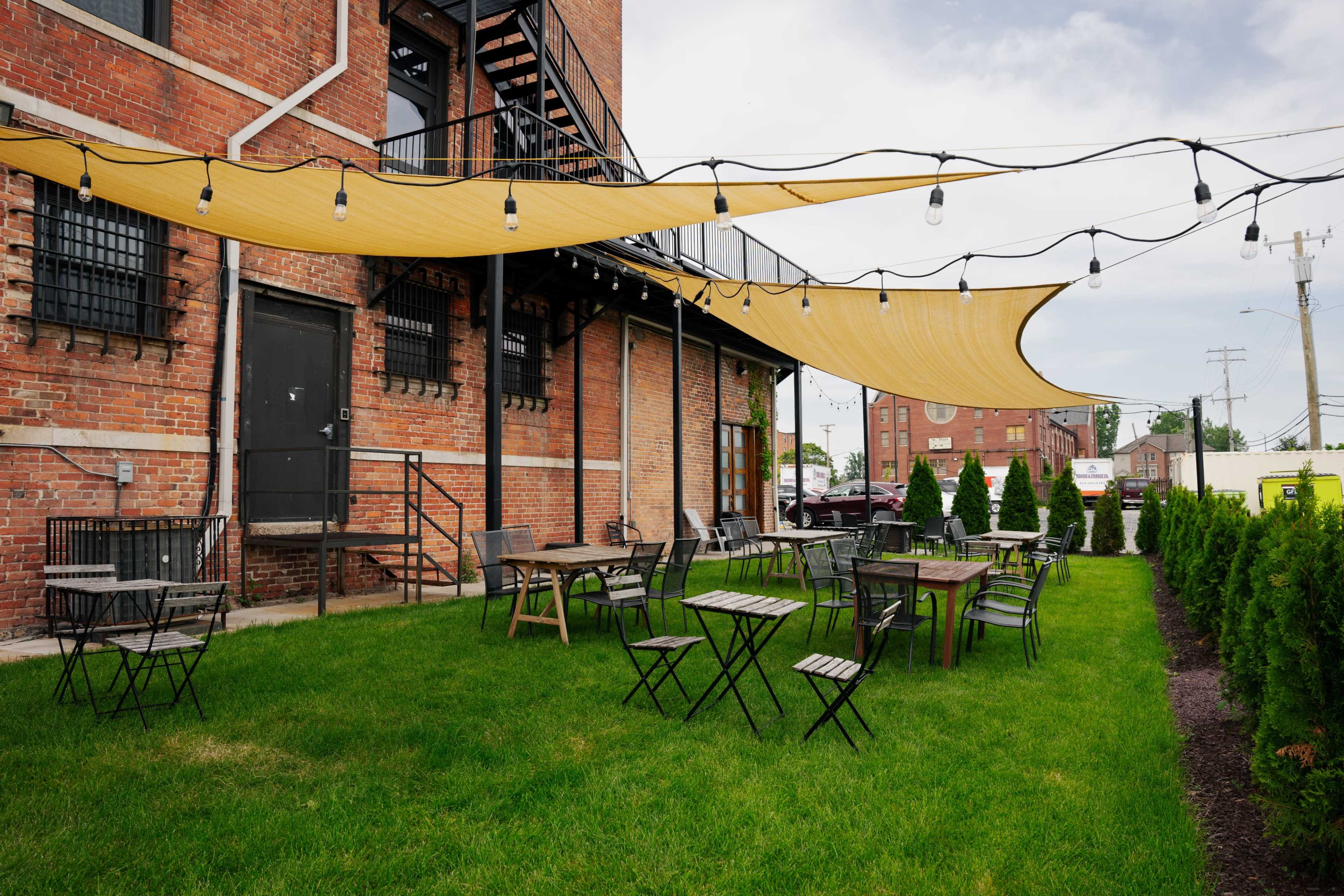 An outdoor patio area with metal tables and chairs beneath yellow shading fabric and string lights, adjacent to a brick building.