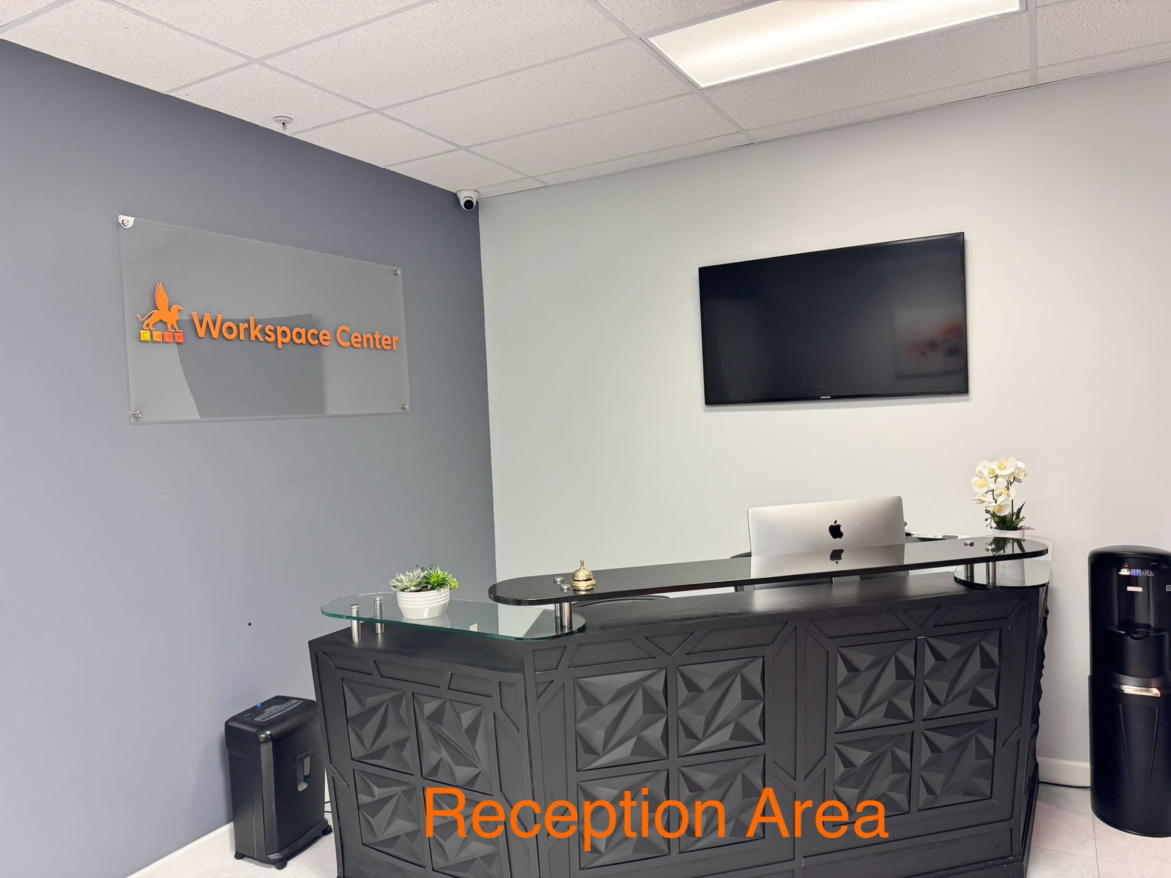 The image shows a modern reception area with a glass-topped desk, a wall-mounted television, and a plant decoration.