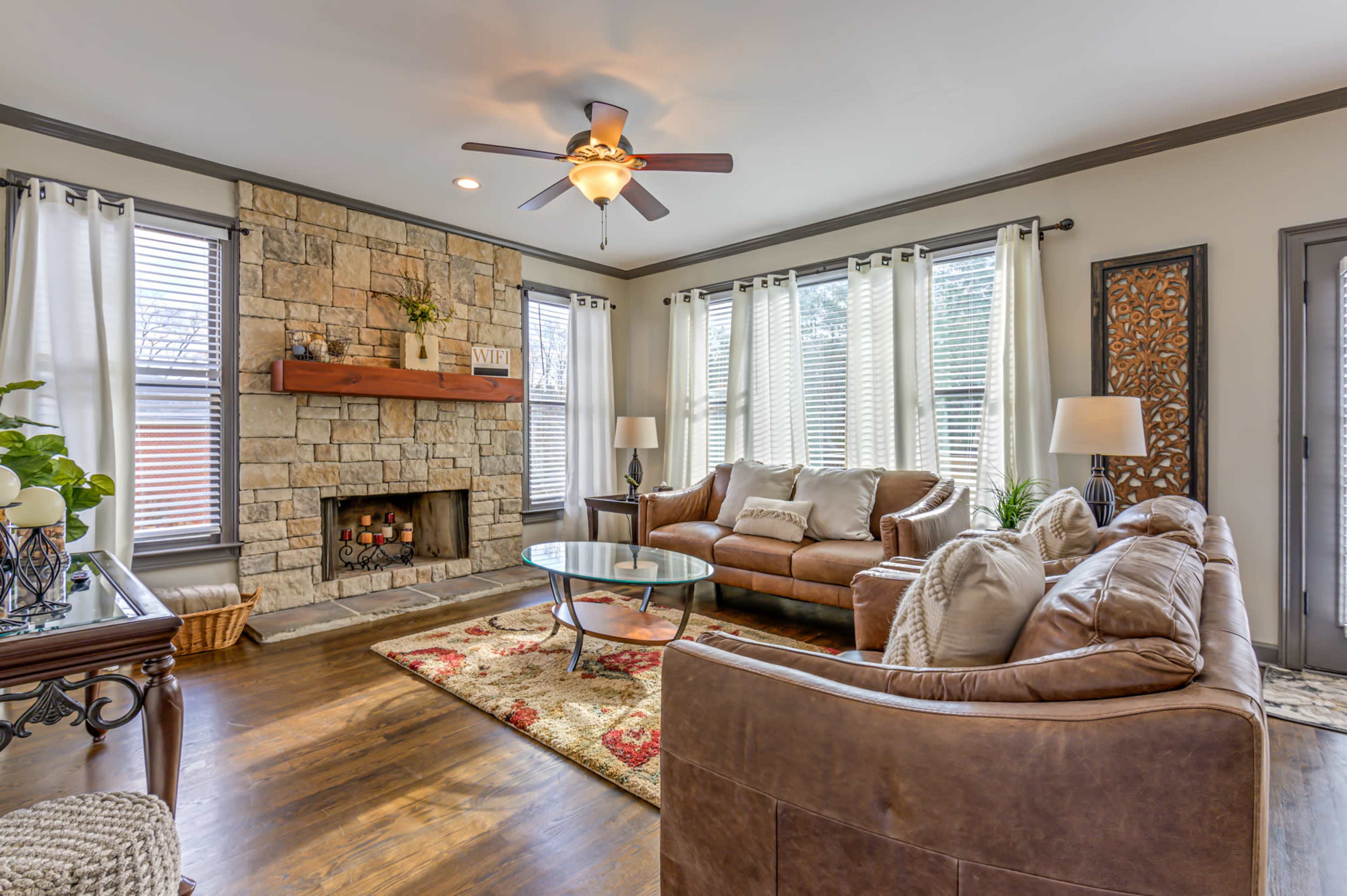 The living room features a stone fireplace, a ceiling fan, and large windows with sheer curtains, surrounded by a combination of leather couches and decorative accents.