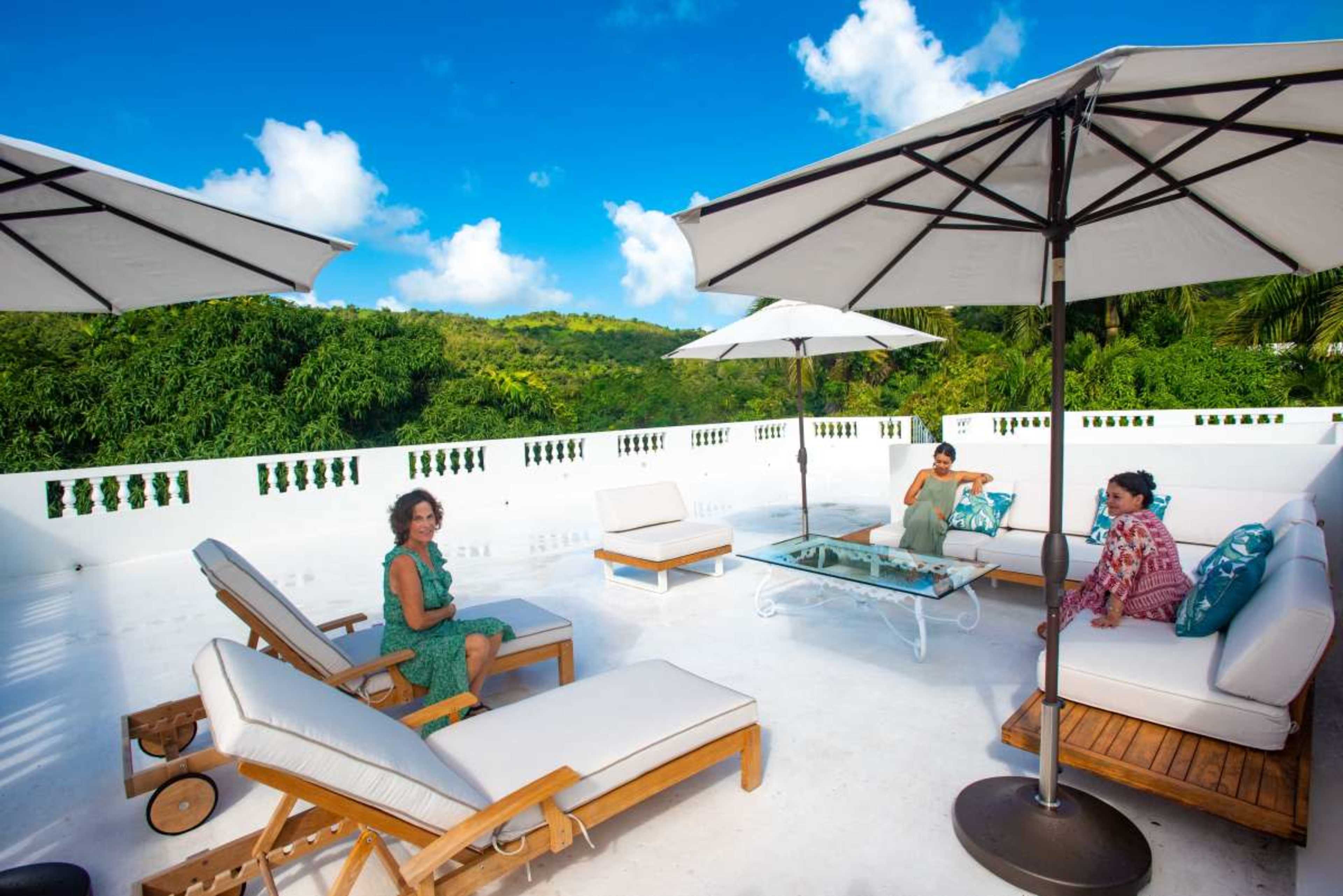 A spacious rooftop terrace features lounge chairs, umbrellas, and a glass table with a scenic view of greenery and blue skies.