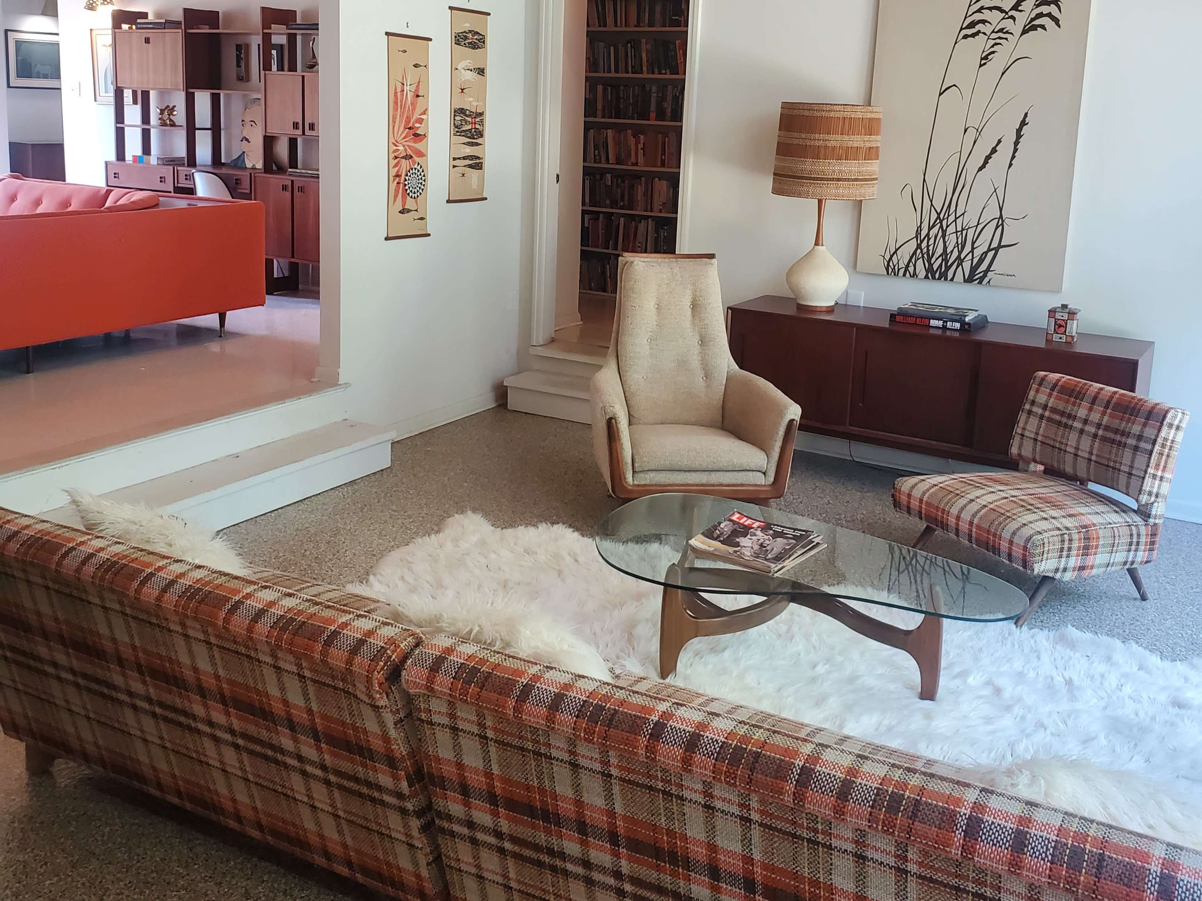 A mid-century modern living room features a plaid sofa, a glass coffee table, and a beige armchair, with a fur rug on a polished floor and bookshelves in the background.