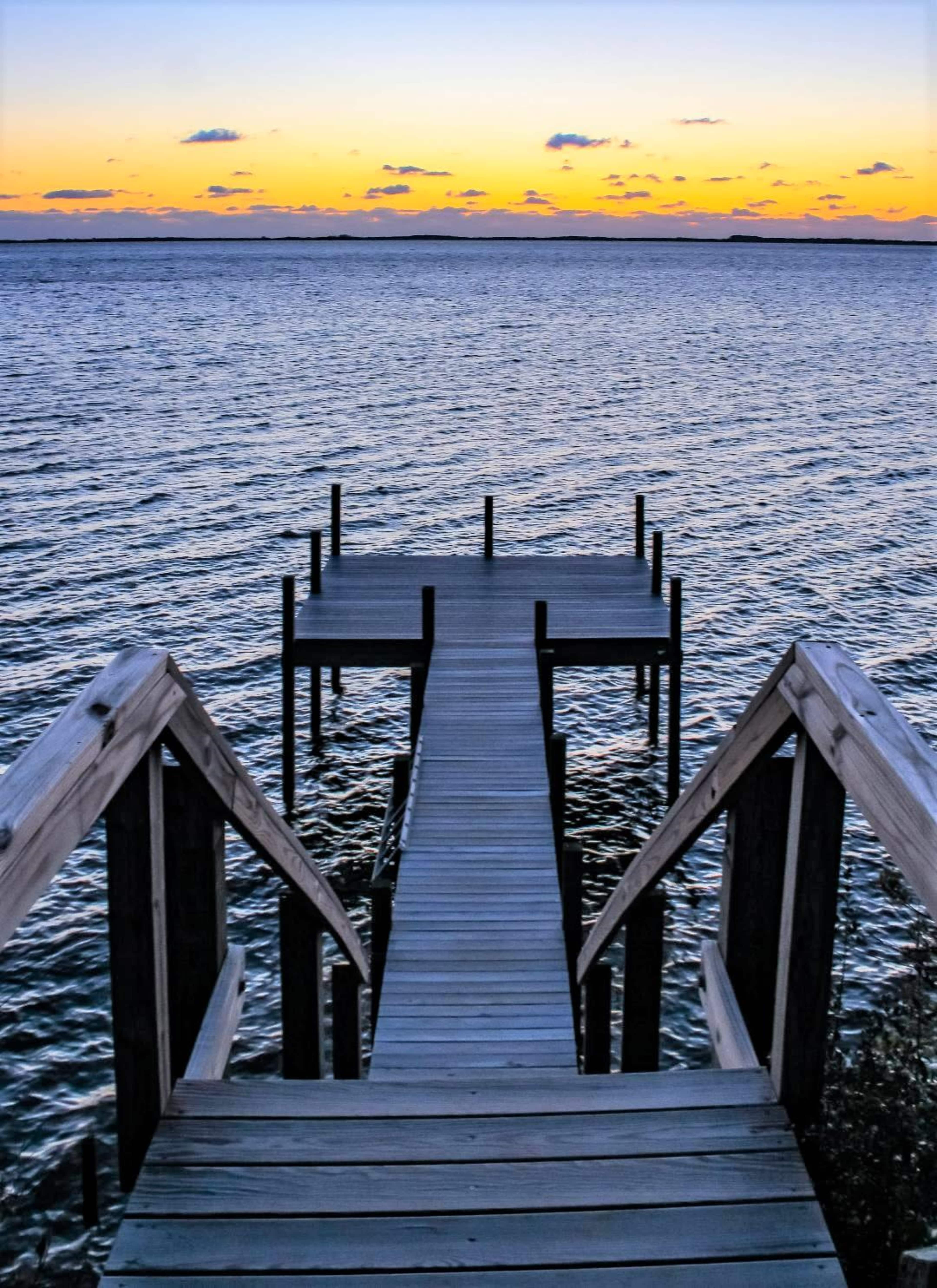 A wooden dock extends over the water toward a sunset sky with colors reflecting on the surface.