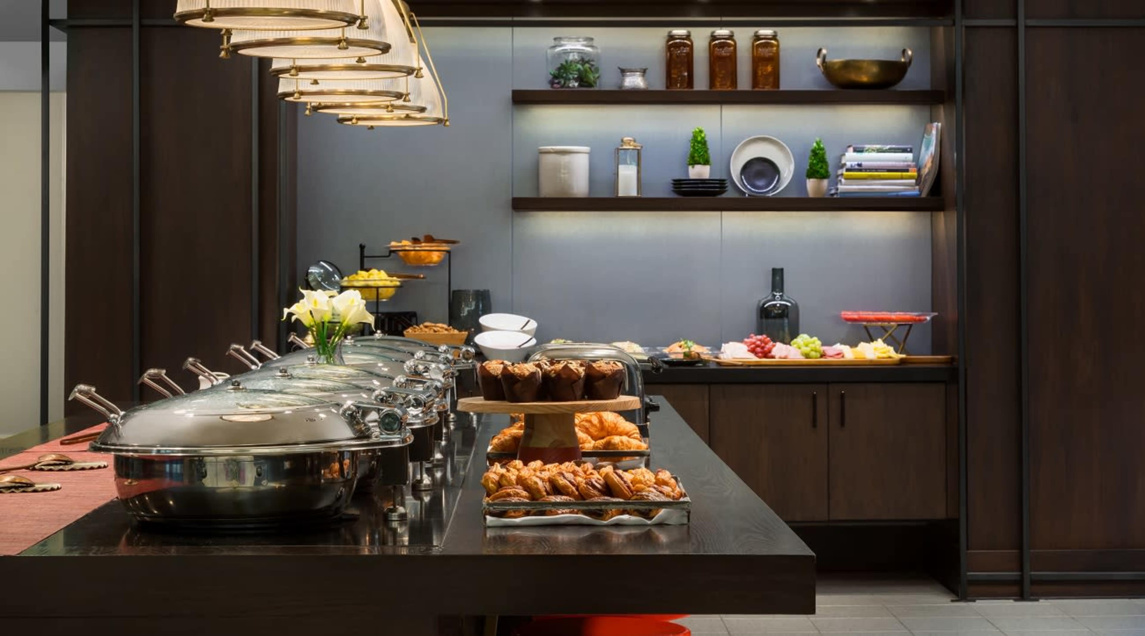 A modern buffet setup features multiple chafing dishes with foods, arranged alongside a display of fruits and pastries on a wooden table in a stylish dining area.
