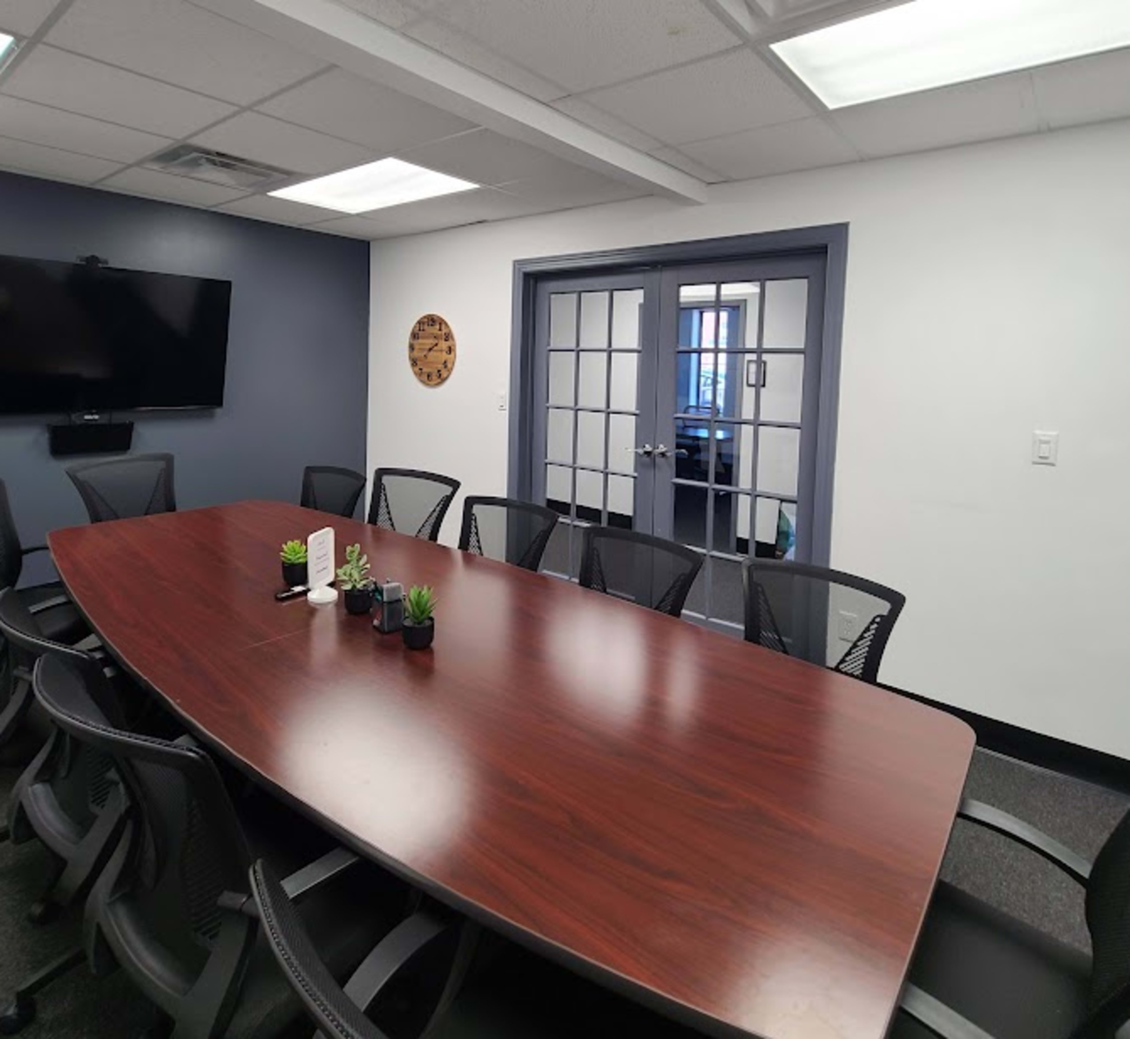 The image shows a conference room with a large wooden table surrounded by black mesh chairs, a wall-mounted TV, and glass doors leading to another room.