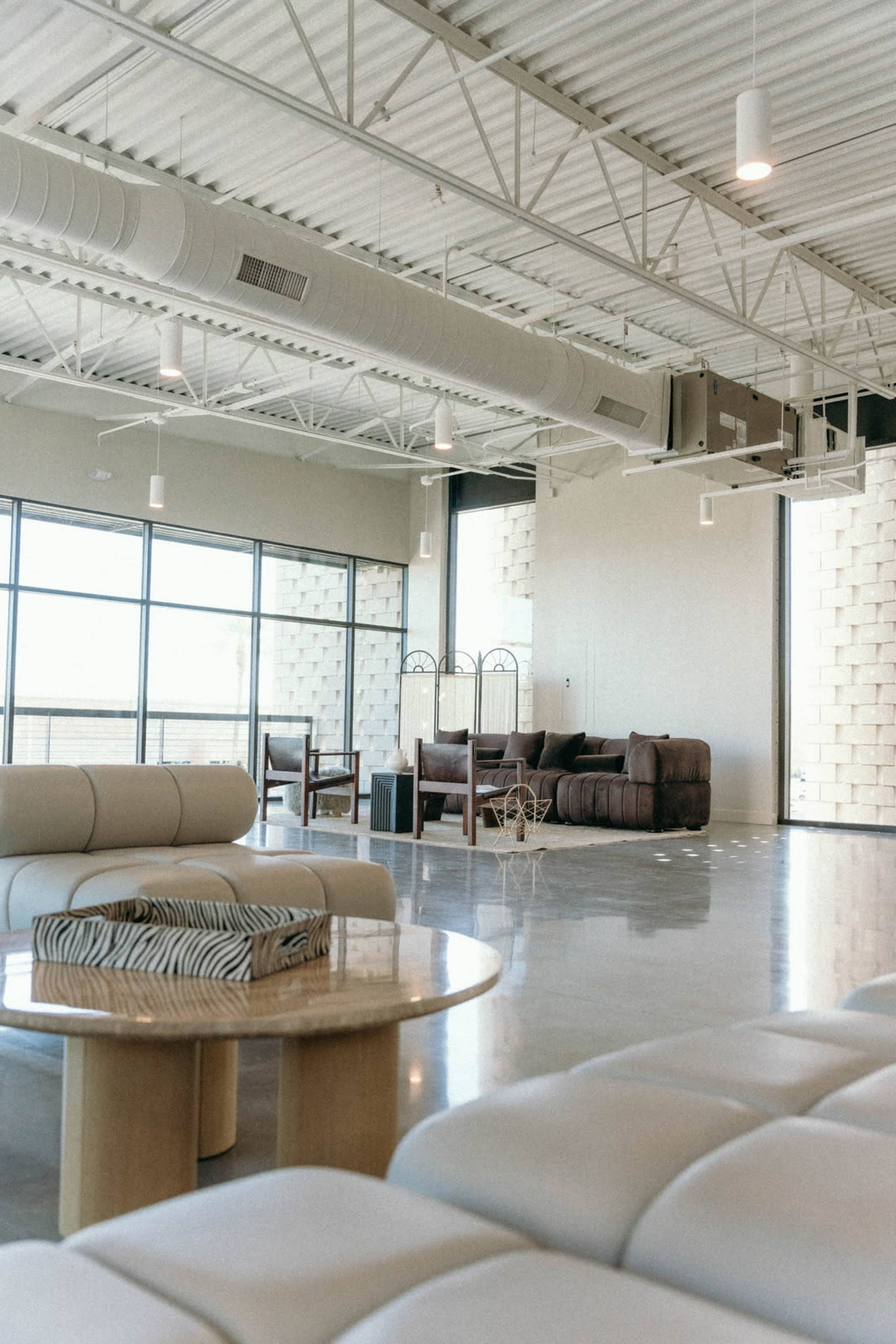 The image shows a modern, open interior space with large windows, featuring minimalist furniture including a circular table and two sofas.