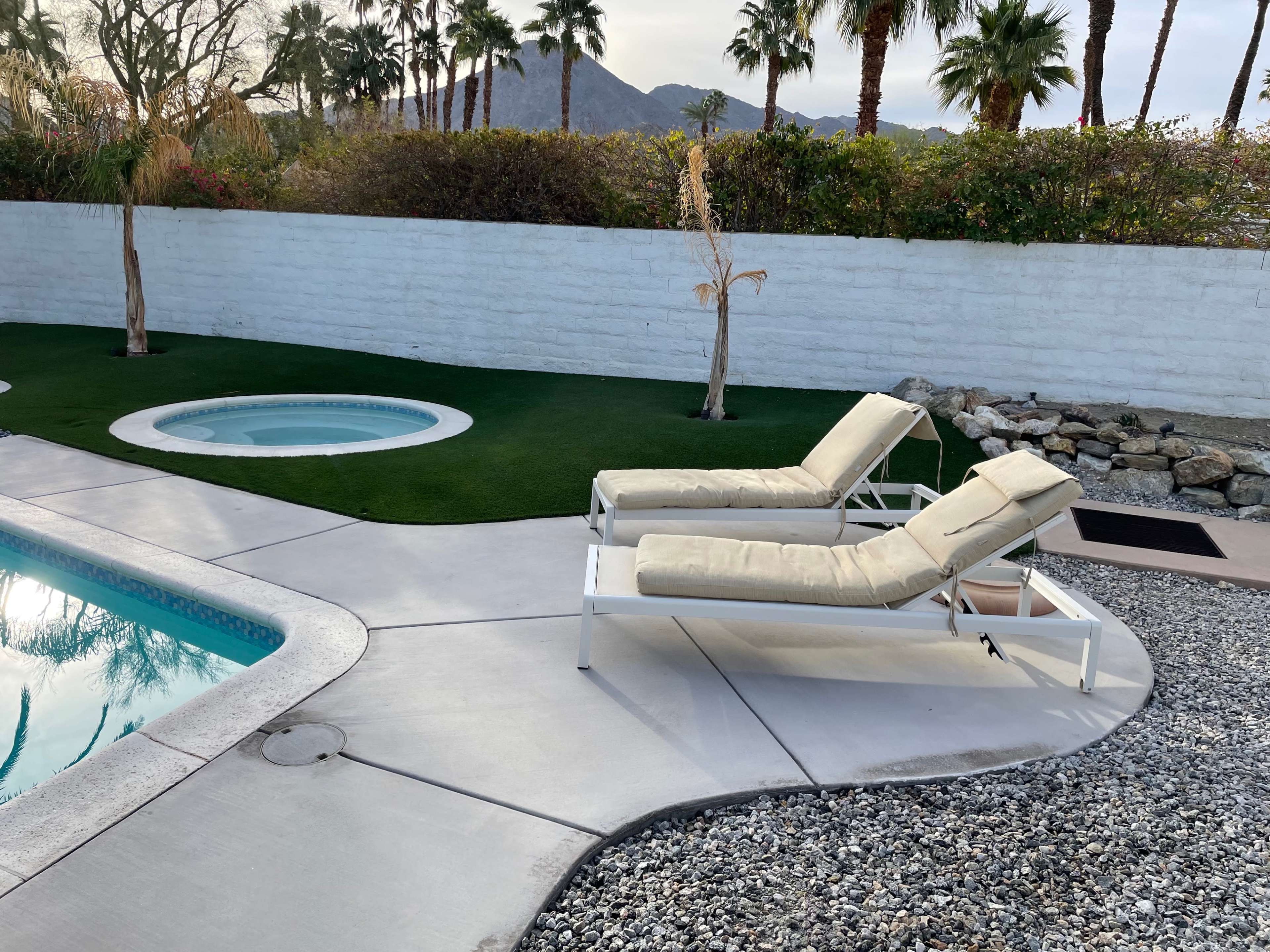 Two adjustable lounge chairs beside a swimming pool and a round hot tub, set against a backdrop of palm trees and a low white wall.