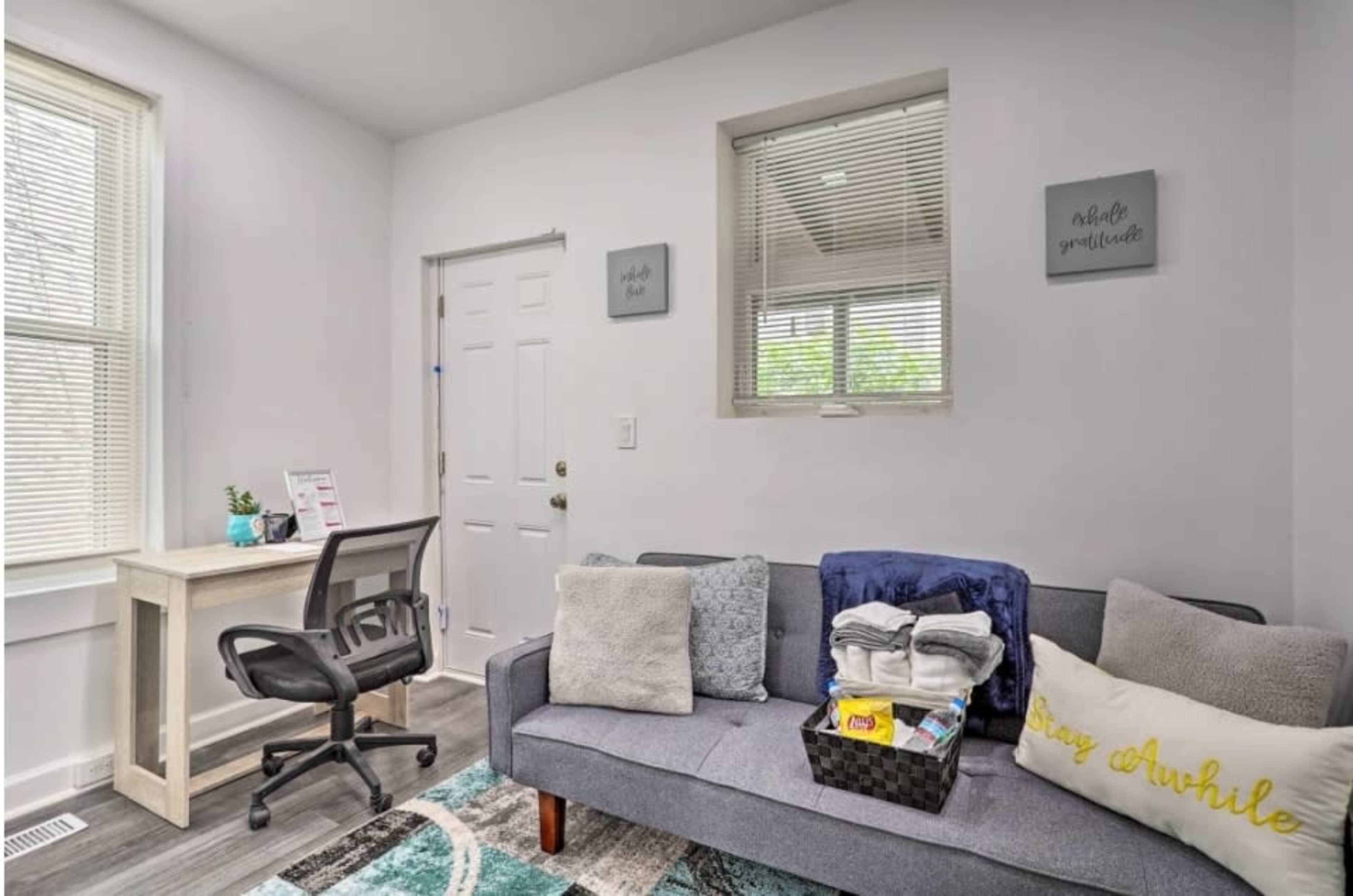 The image shows a small room featuring a gray couch with throw pillows, a desk with an office chair, and a door leading outside.