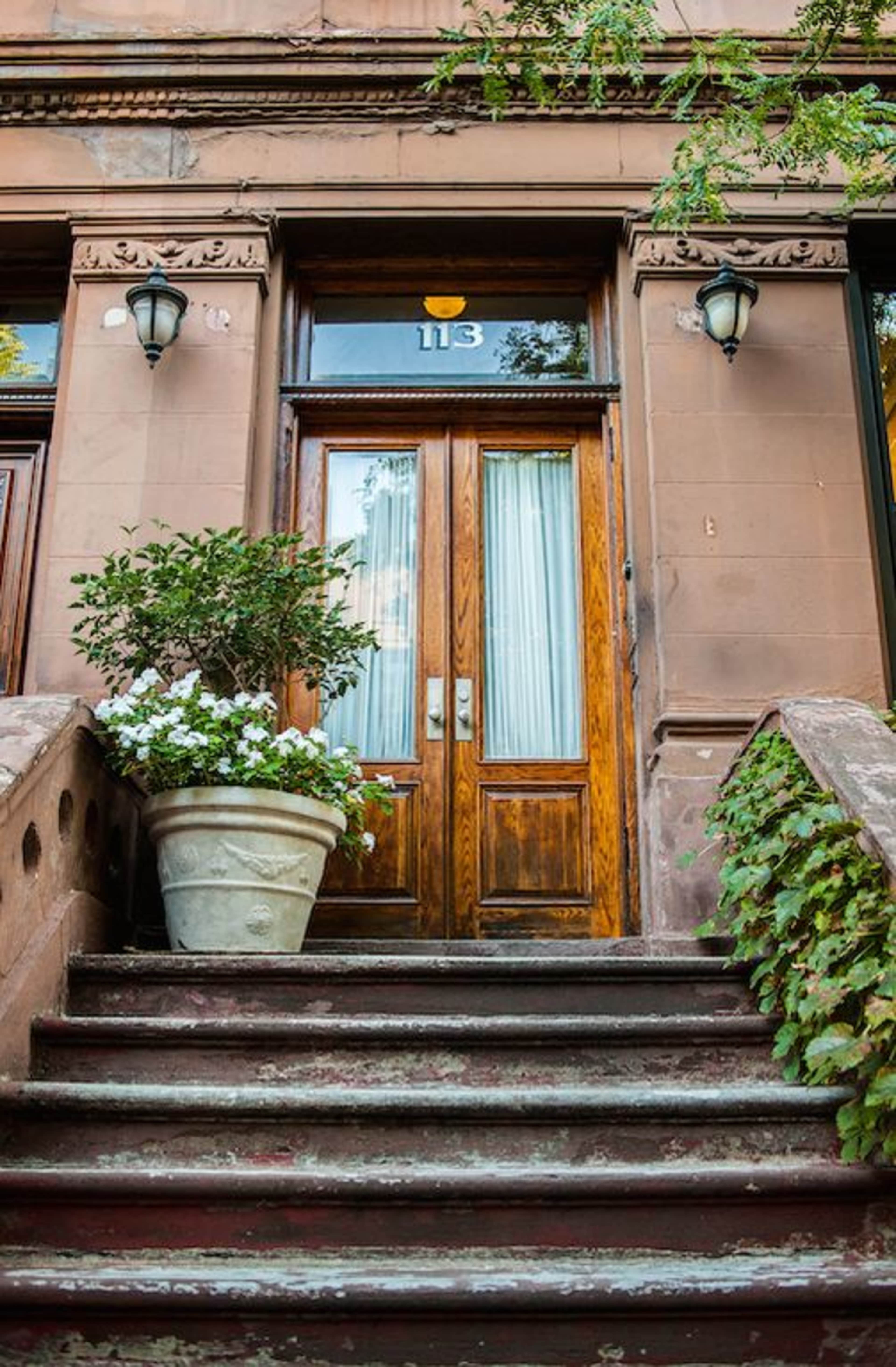 The image shows a pair of wooden doors with glass panes, flanked by decorative light fixtures, beneath a set of steps that includes a potted plant with white flowers.