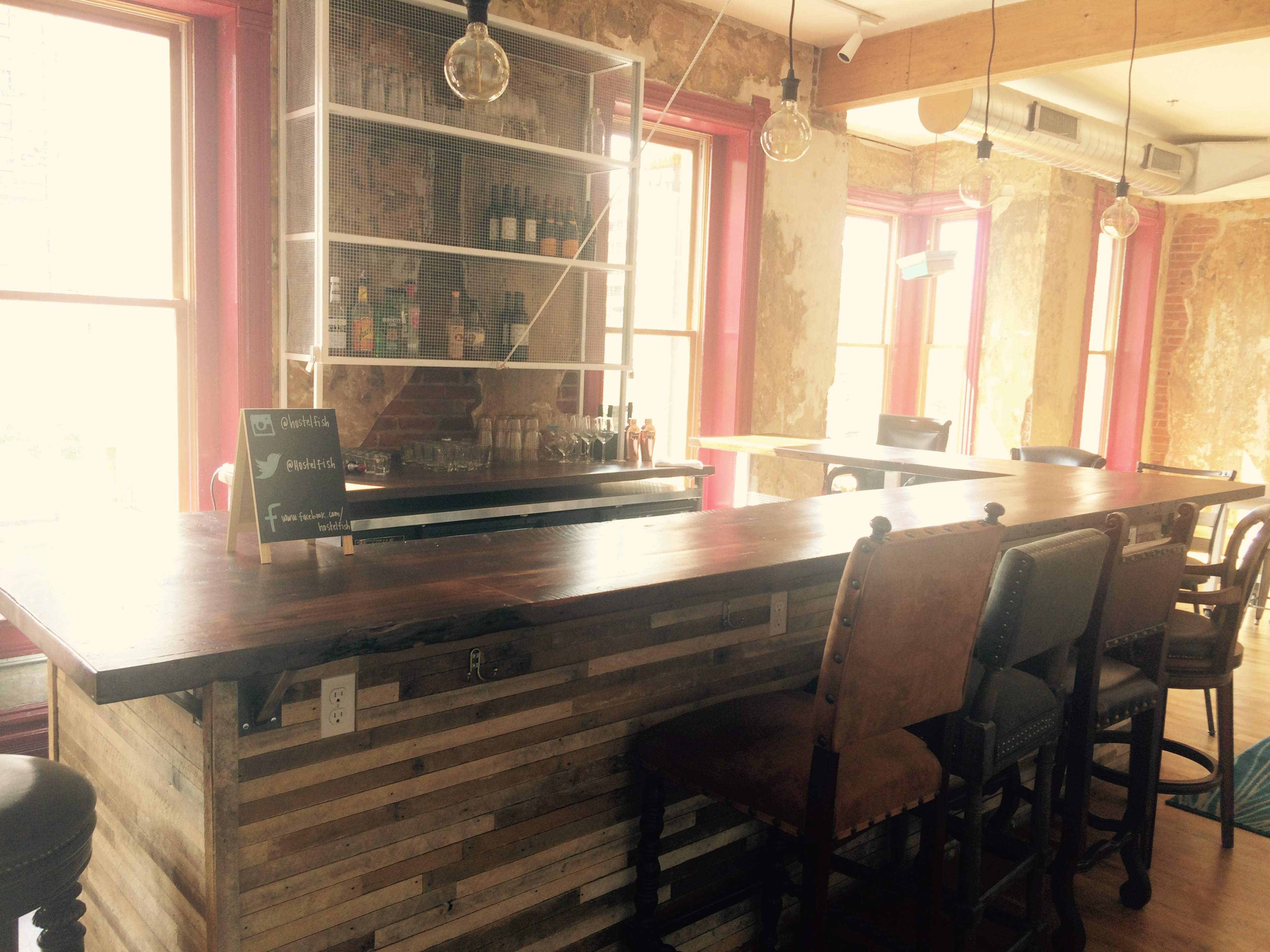 A rustic bar area with a wooden counter, several high-backed chairs, and a display of bottles in a well-lit interior with large windows.