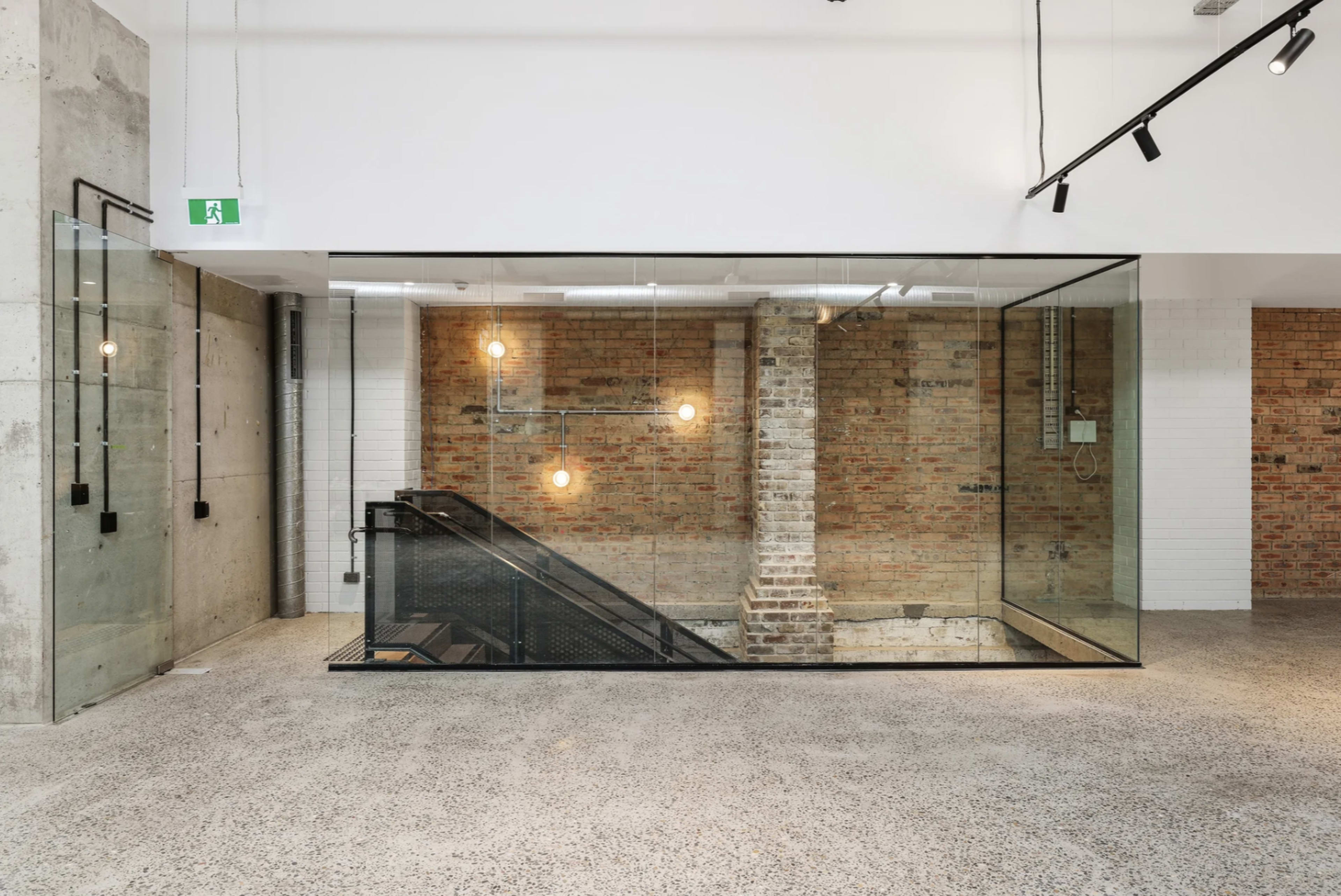 A modern interior space featuring a glass-walled staircase that leads to a lower level, with exposed brick walls and overhead lighting.