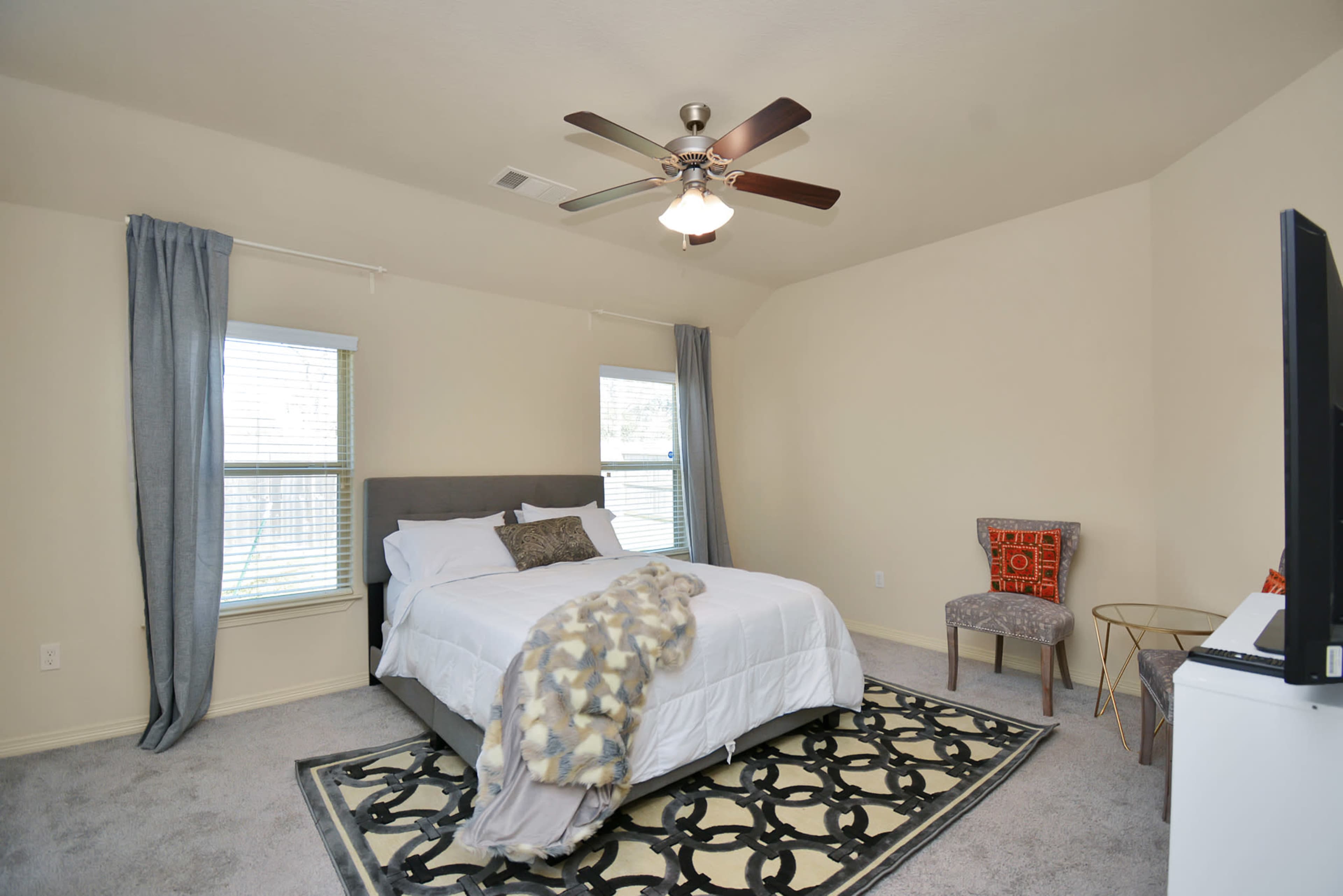 A bedroom with a queen-sized bed, a ceiling fan, two windows, a chair in the corner, and a decorative rug on the floor.