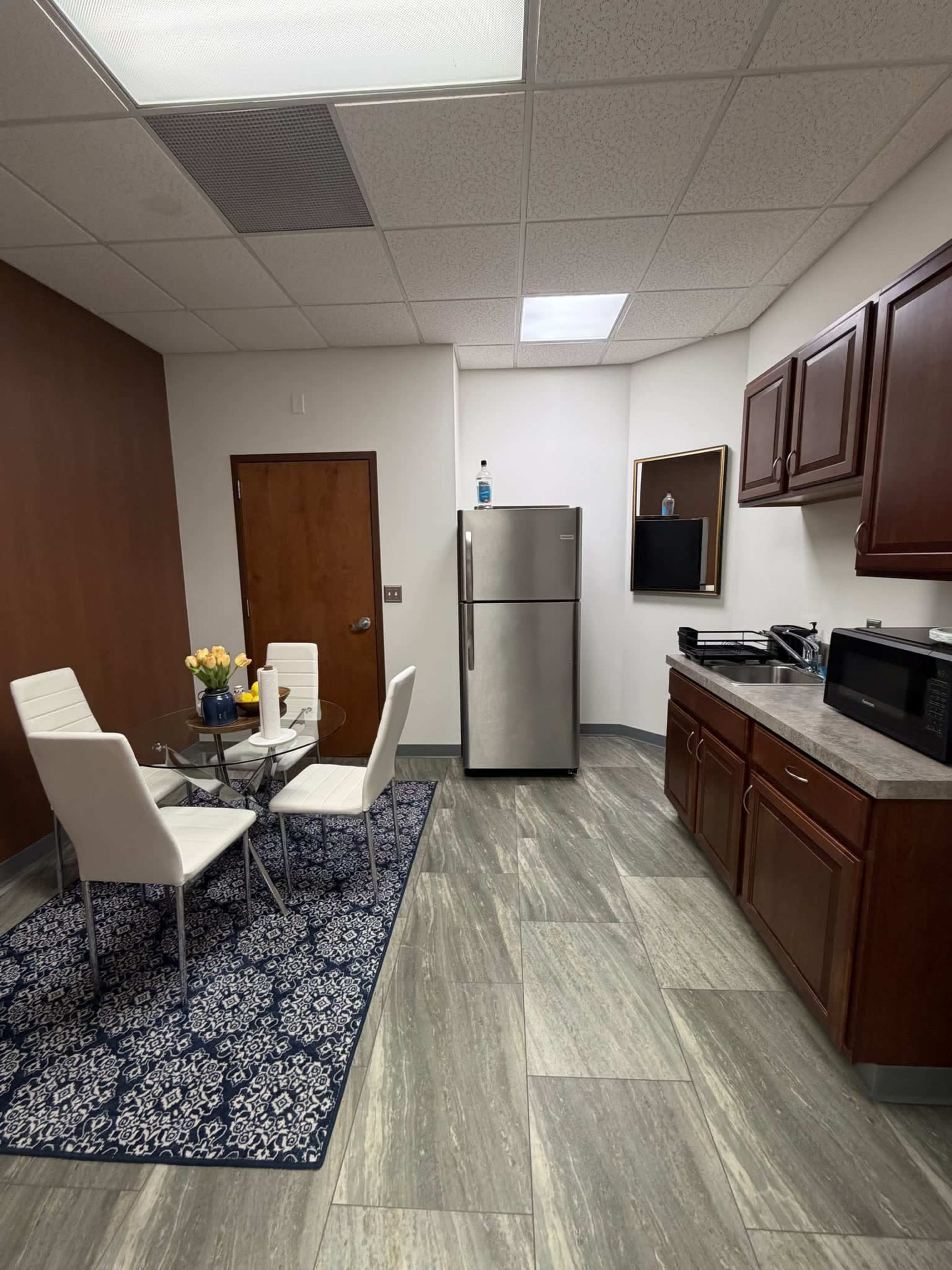 A small break room features a round table with two white chairs, a refrigerator, a microwave, and dark wood cabinets against a wall.