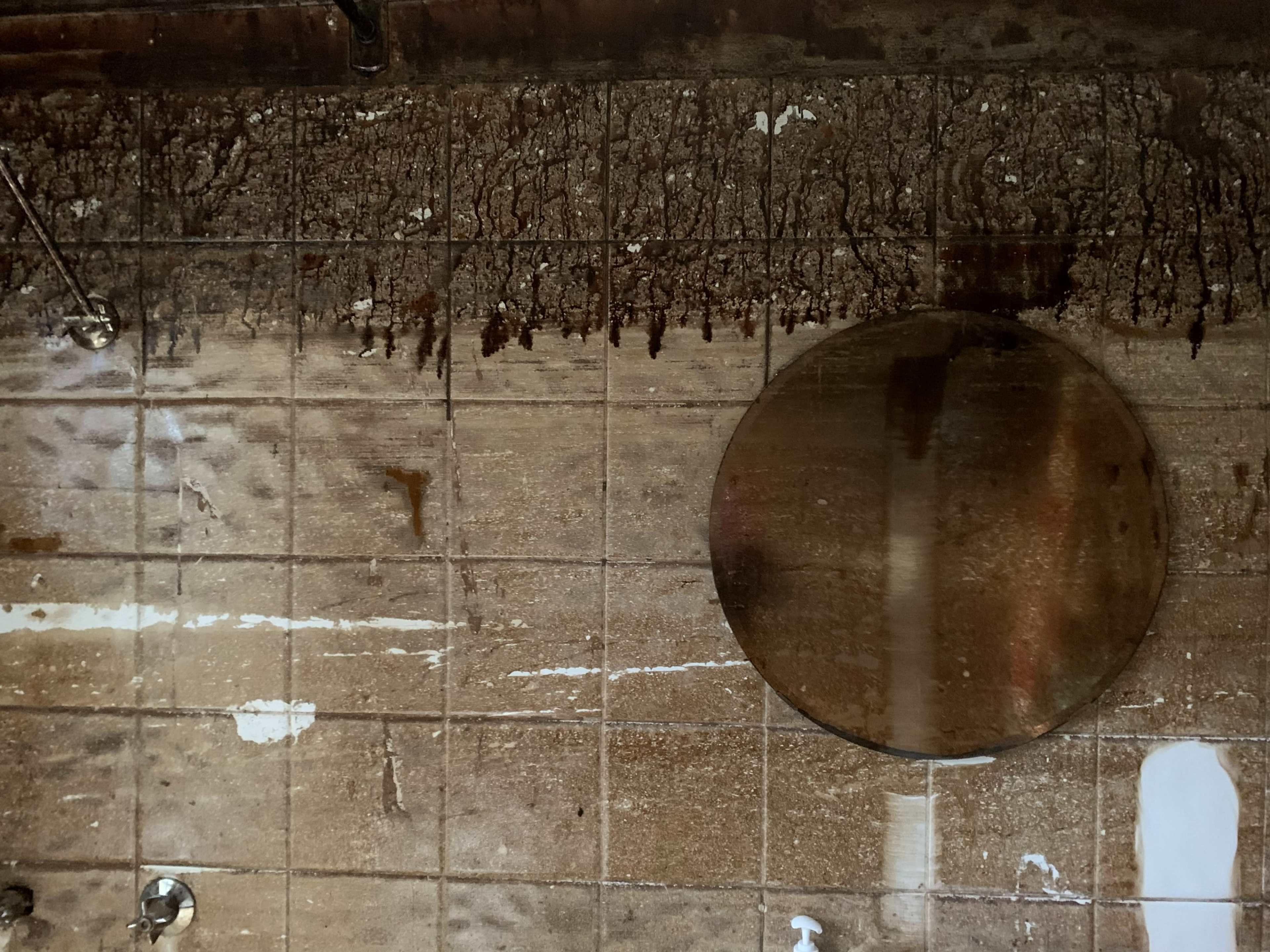 The image shows a tiled wall with streaks of water damage and a circular mirror mounted on it.