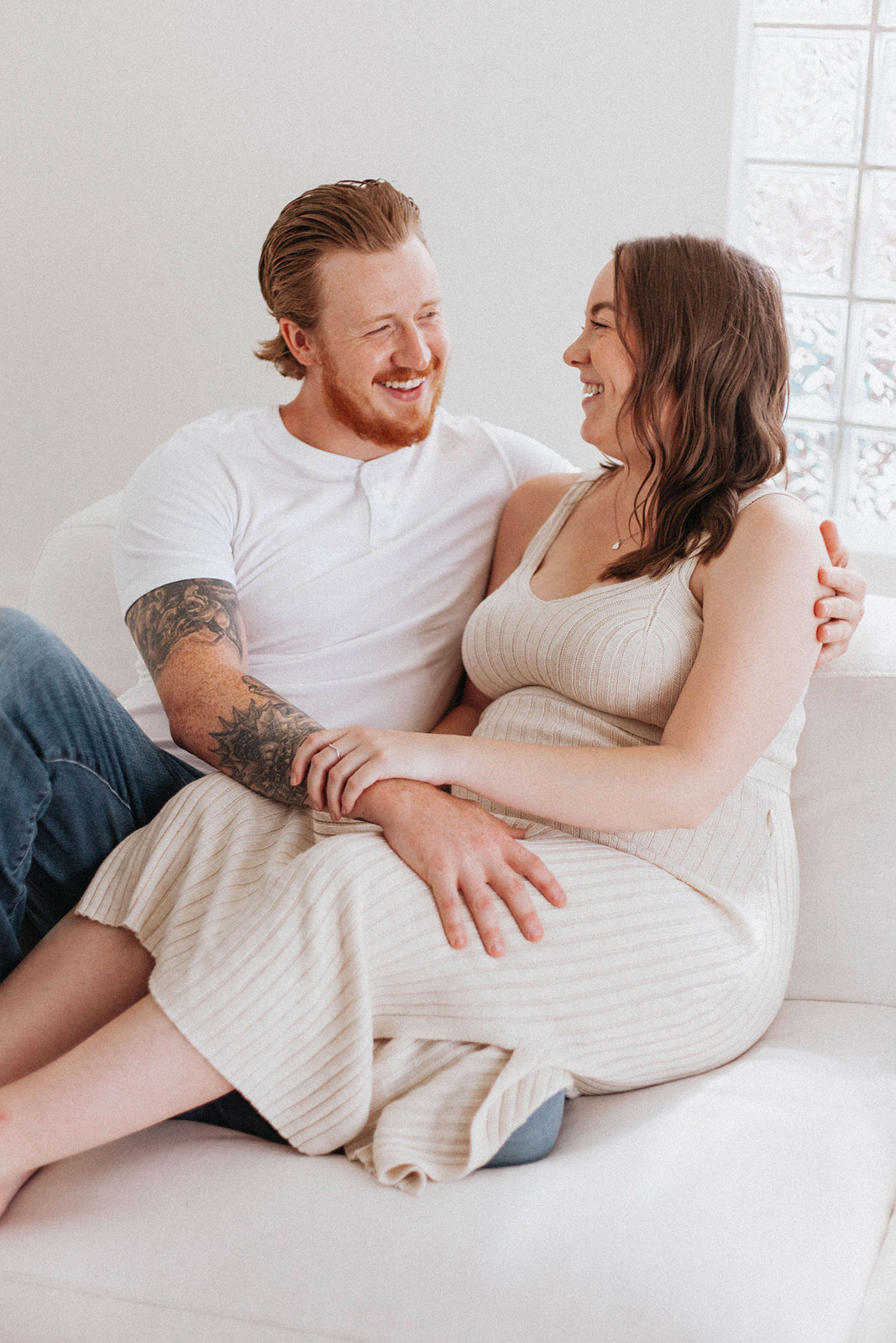 A minimalist couple poses for a maternity photo shoot on a white couch.