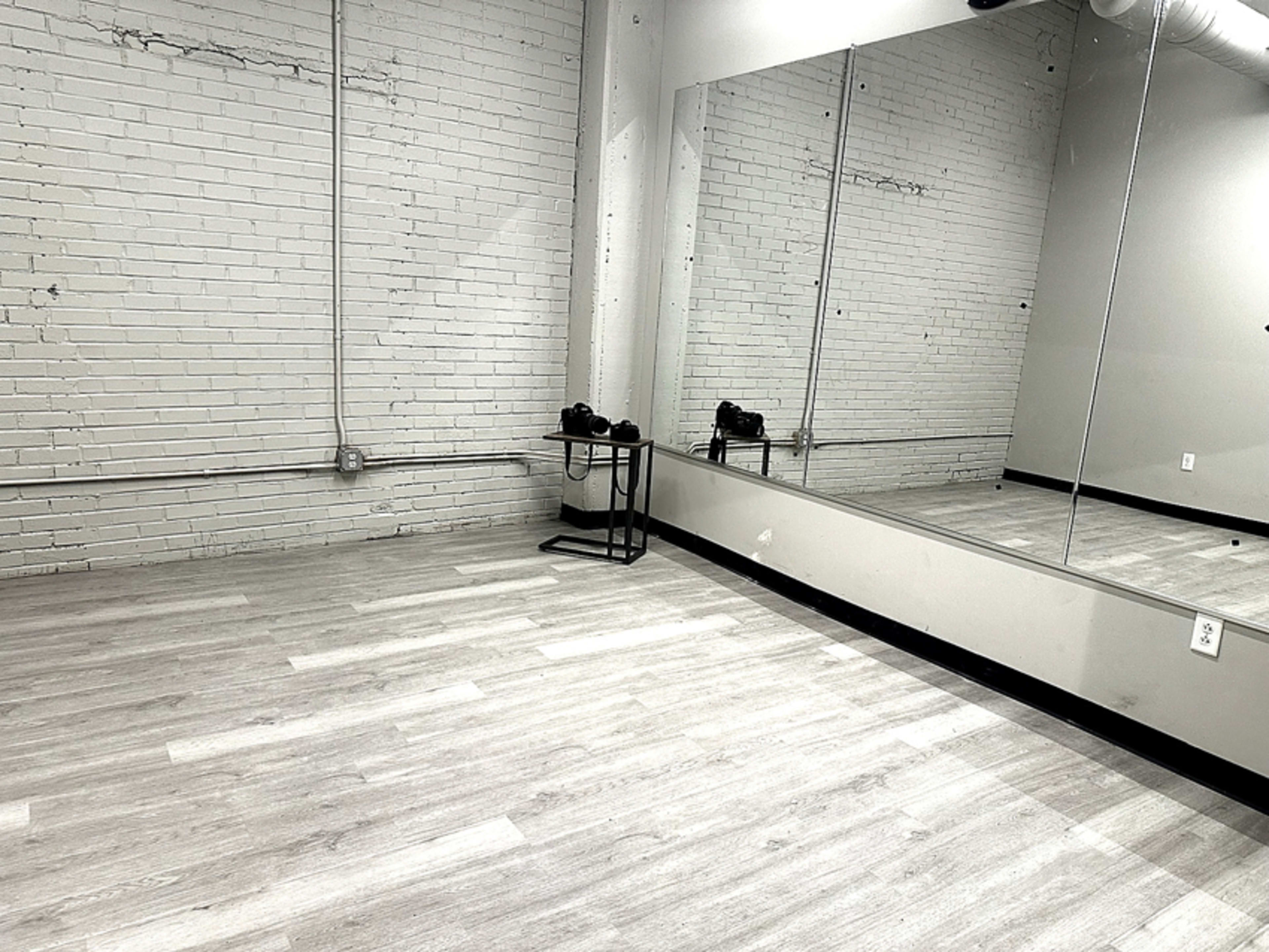 The image shows an empty room with light-colored wooden flooring, exposed white brick walls, and a large mirror along one side.