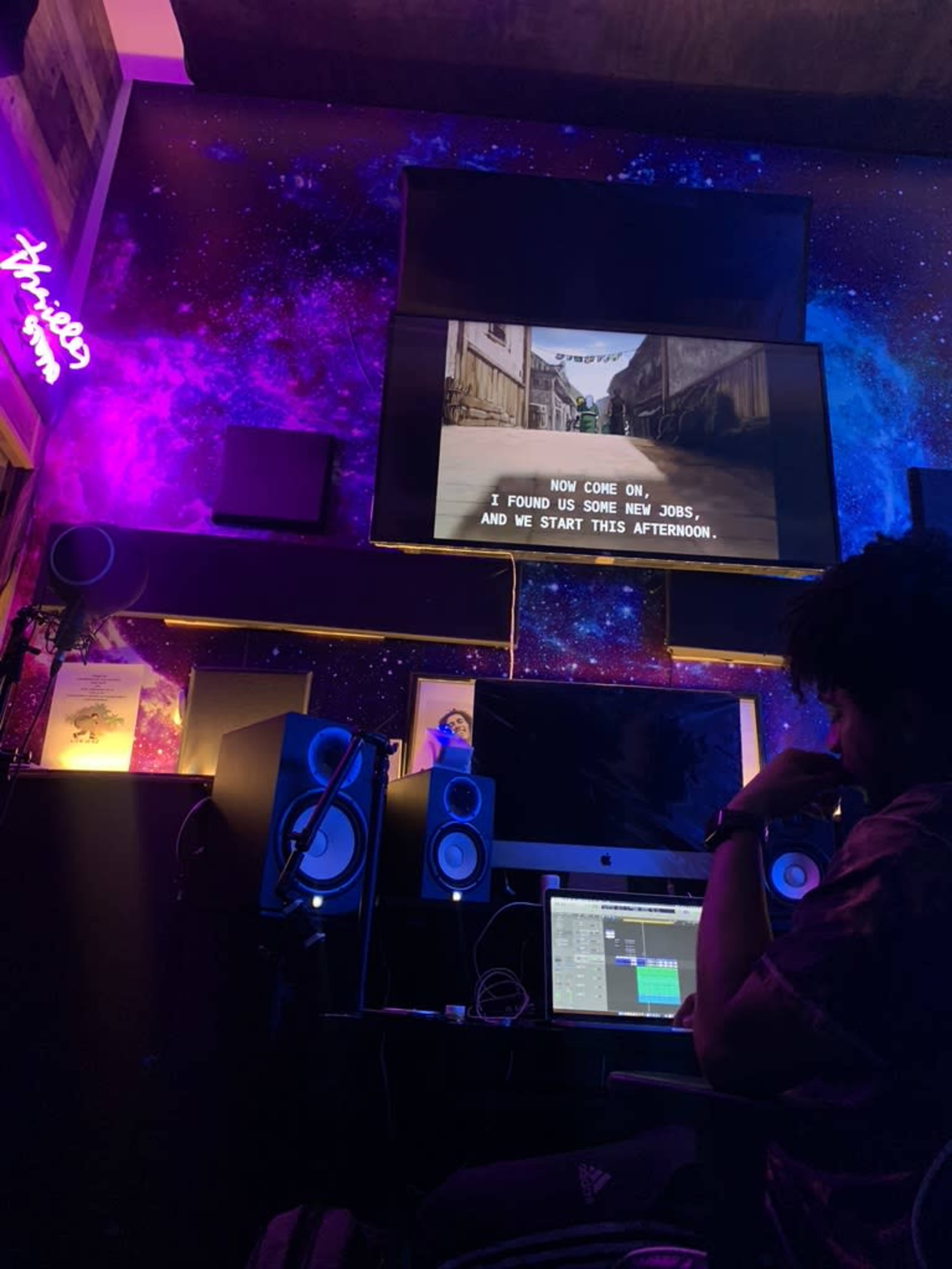 A person sits in a dimly lit music studio, working on a computer with speakers, while subtitles display on a screen above.