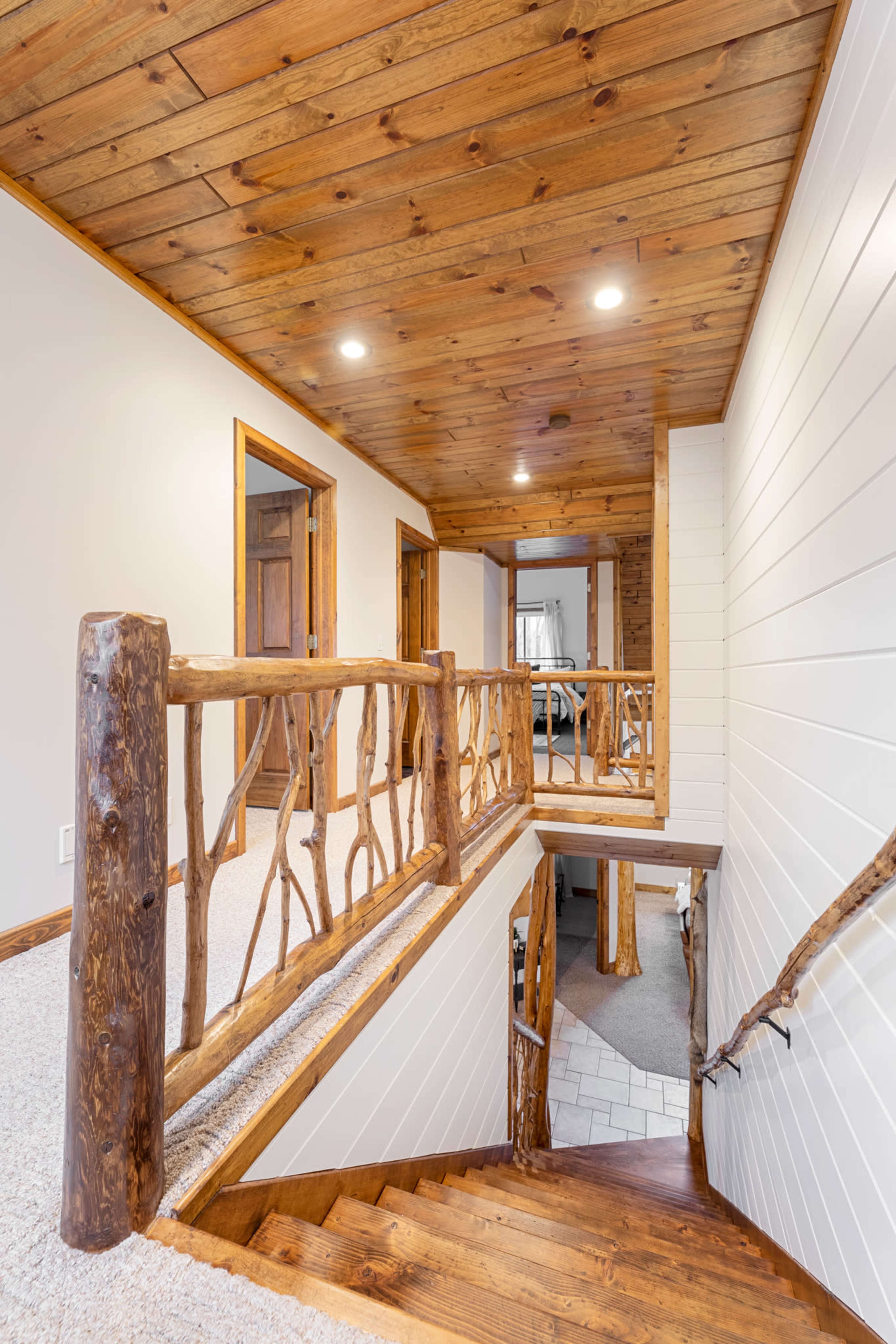 The image shows a wooden staircase with a rustic railing leading down to a lower level, set in a well-lit hall featuring wood-paneled walls and ceiling.