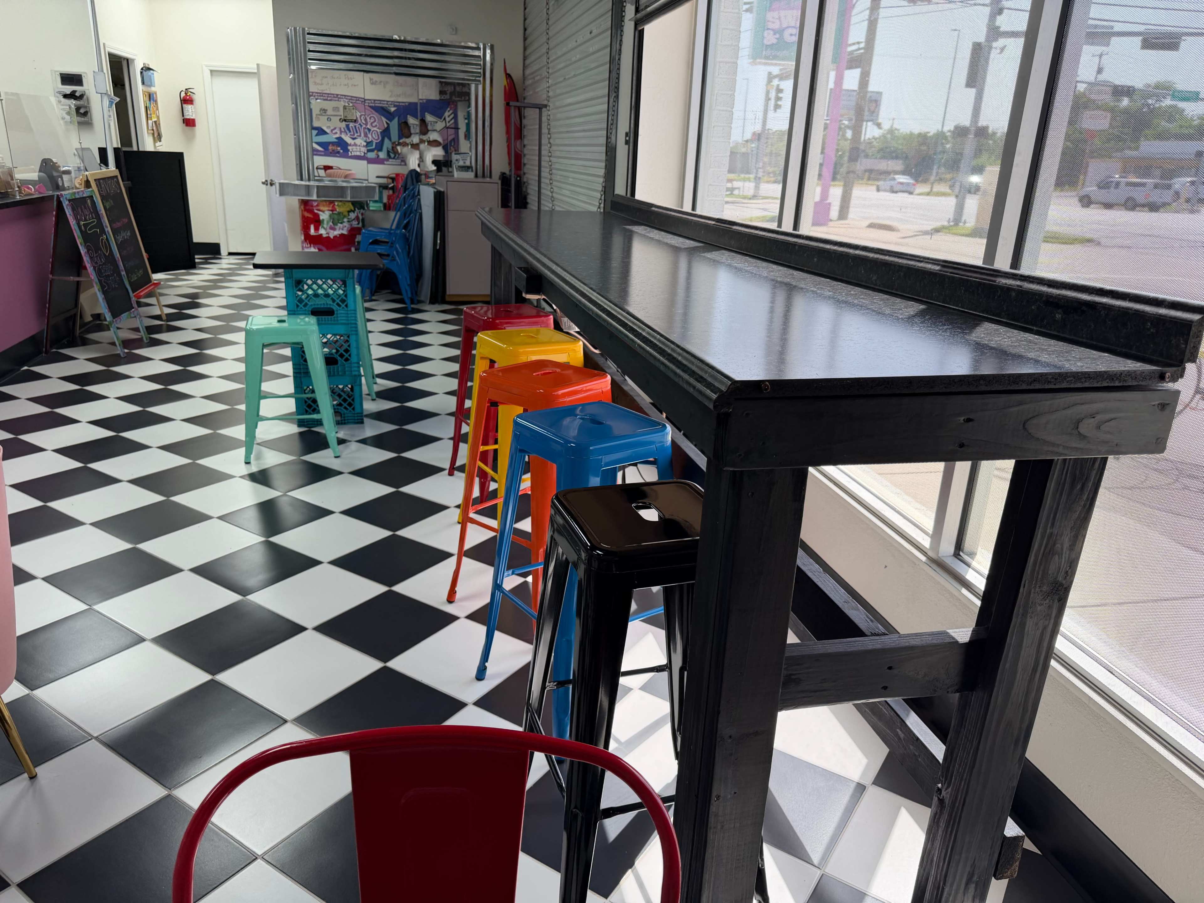 The image shows a brightly colored café interior featuring a long black table with colorful stools arranged along one side, set against a checkered floor.
