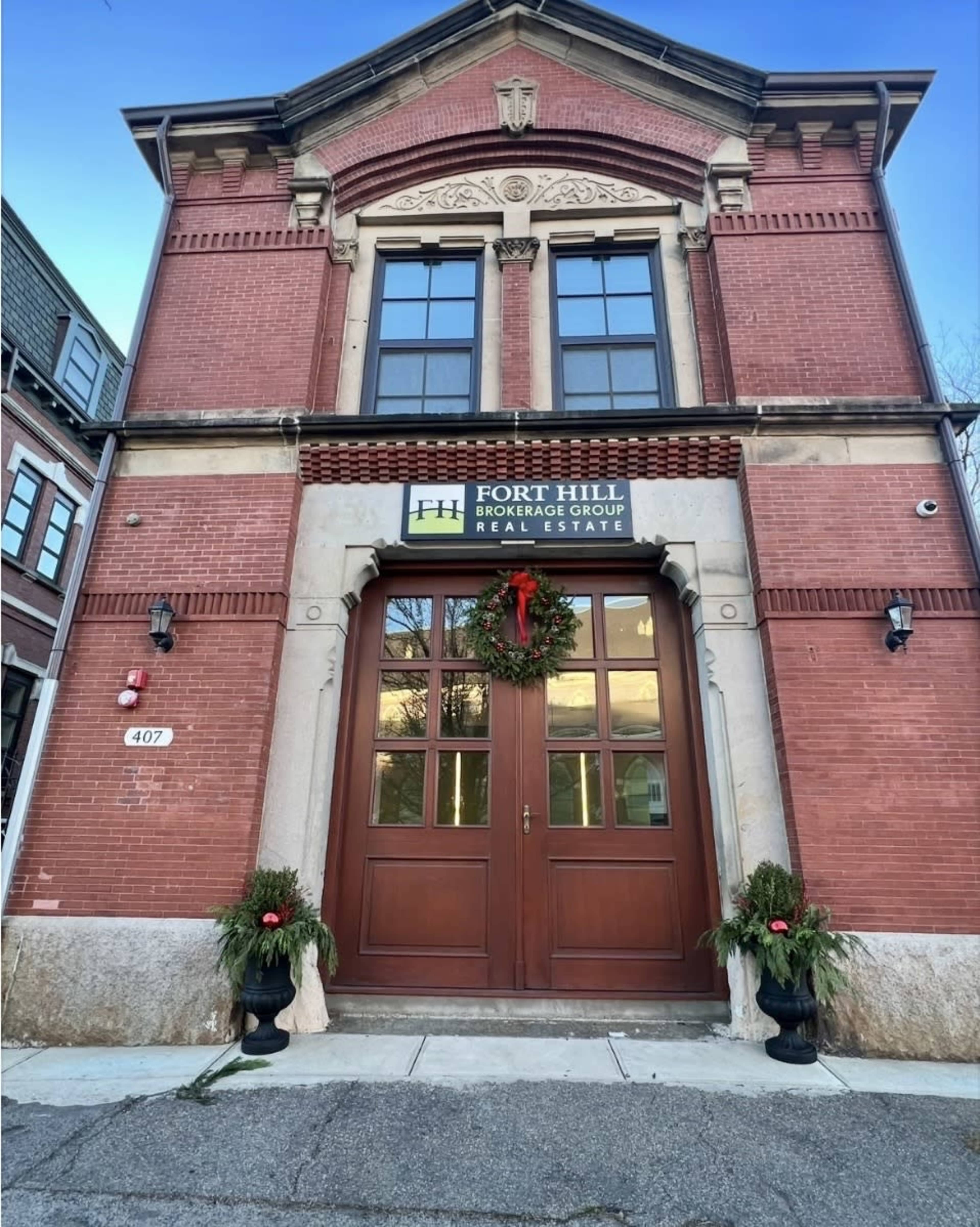 The image shows a brick building with a large wooden door, flanked by potted plants, and a sign indicating it is the Fort Hill Brokerage Group for real estate.
