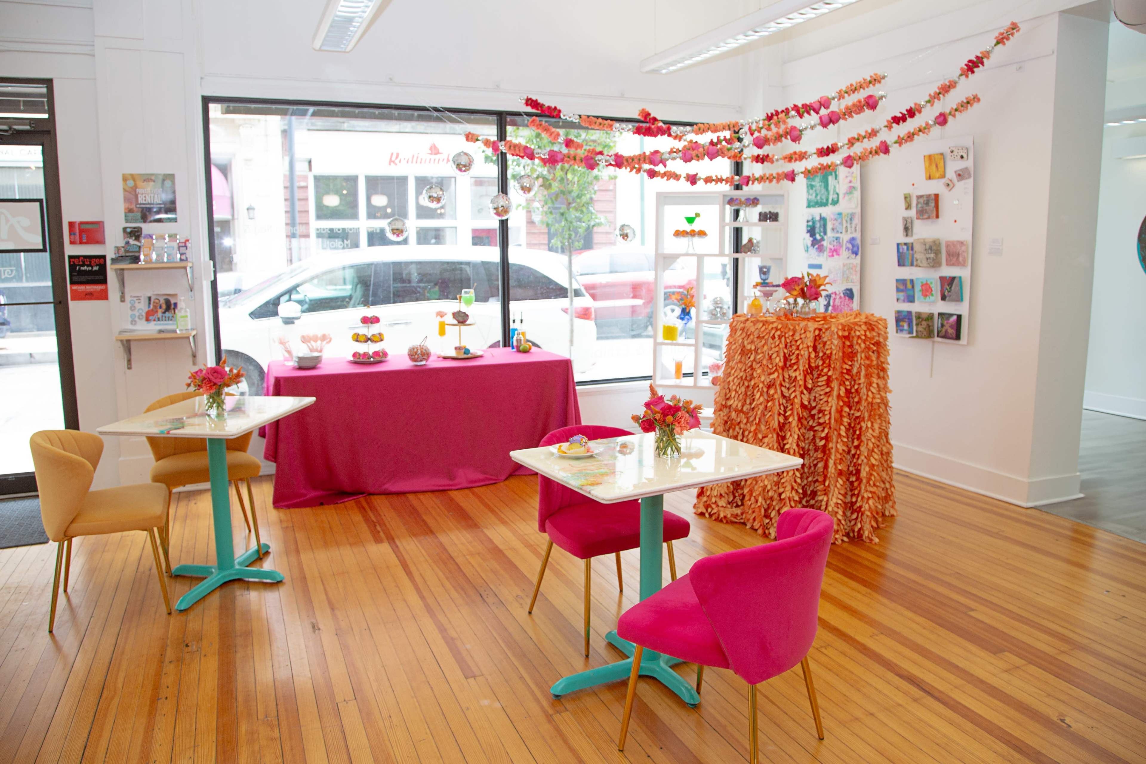 An art space with tables decorated with flowers, a pink tablecloth, and colorful decorations hanging from the ceiling.