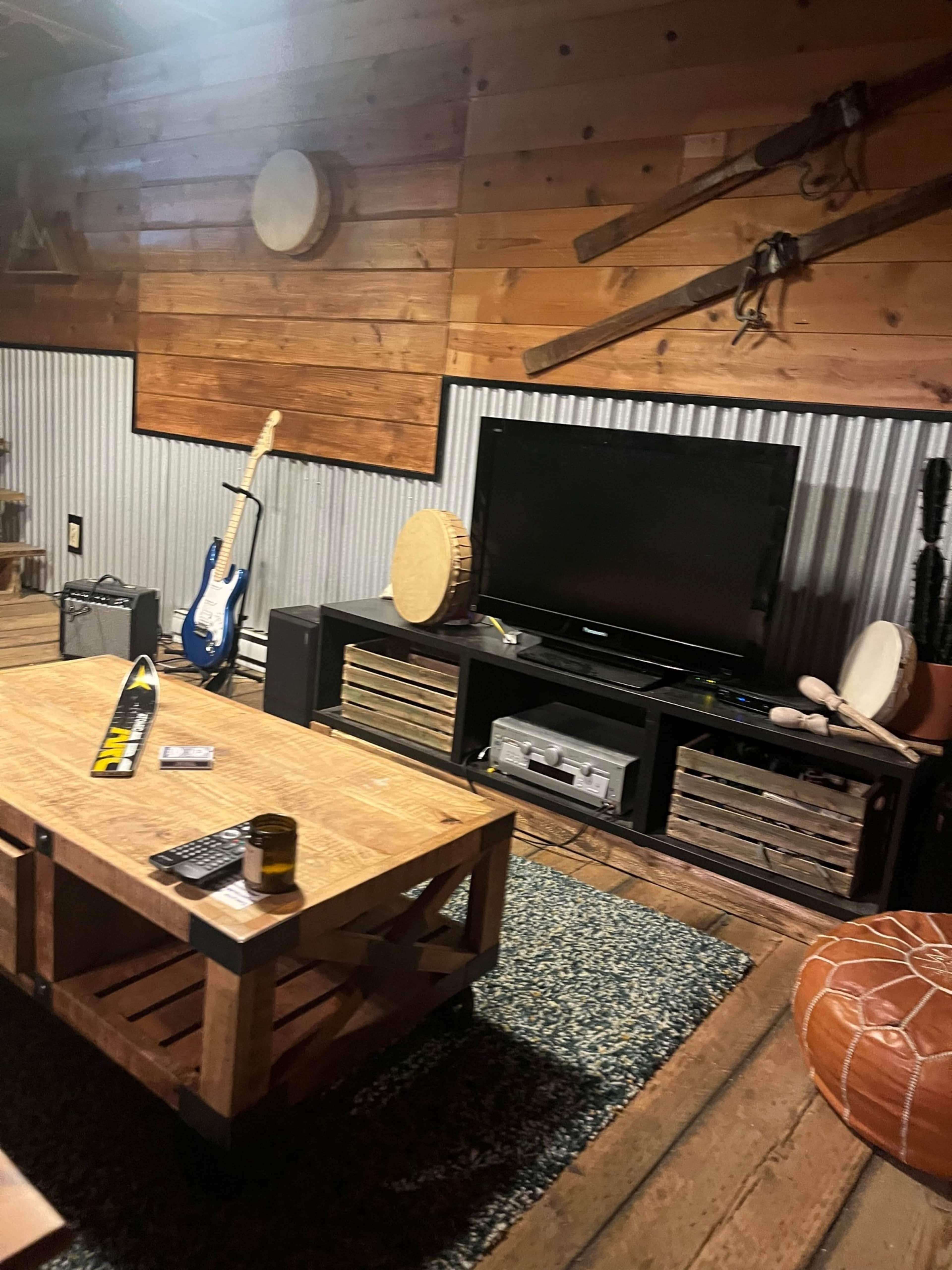 The room features a rustic design with wooden walls, a guitar leaning against the wall, a TV on a low black cabinet, and a wooden coffee table.