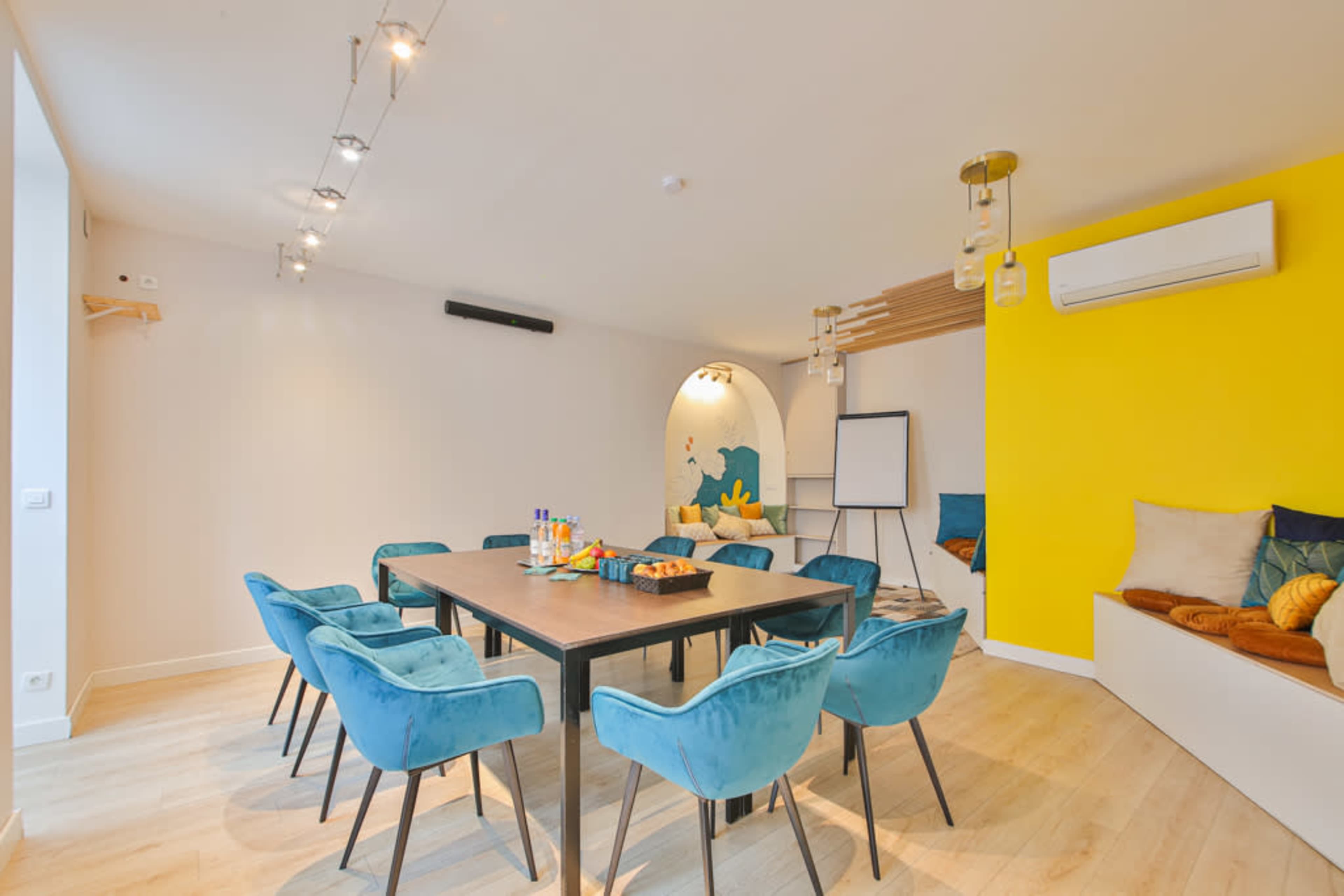 A modern meeting room features a large table surrounded by blue chairs, a whiteboard, and bright yellow accents on the wall.