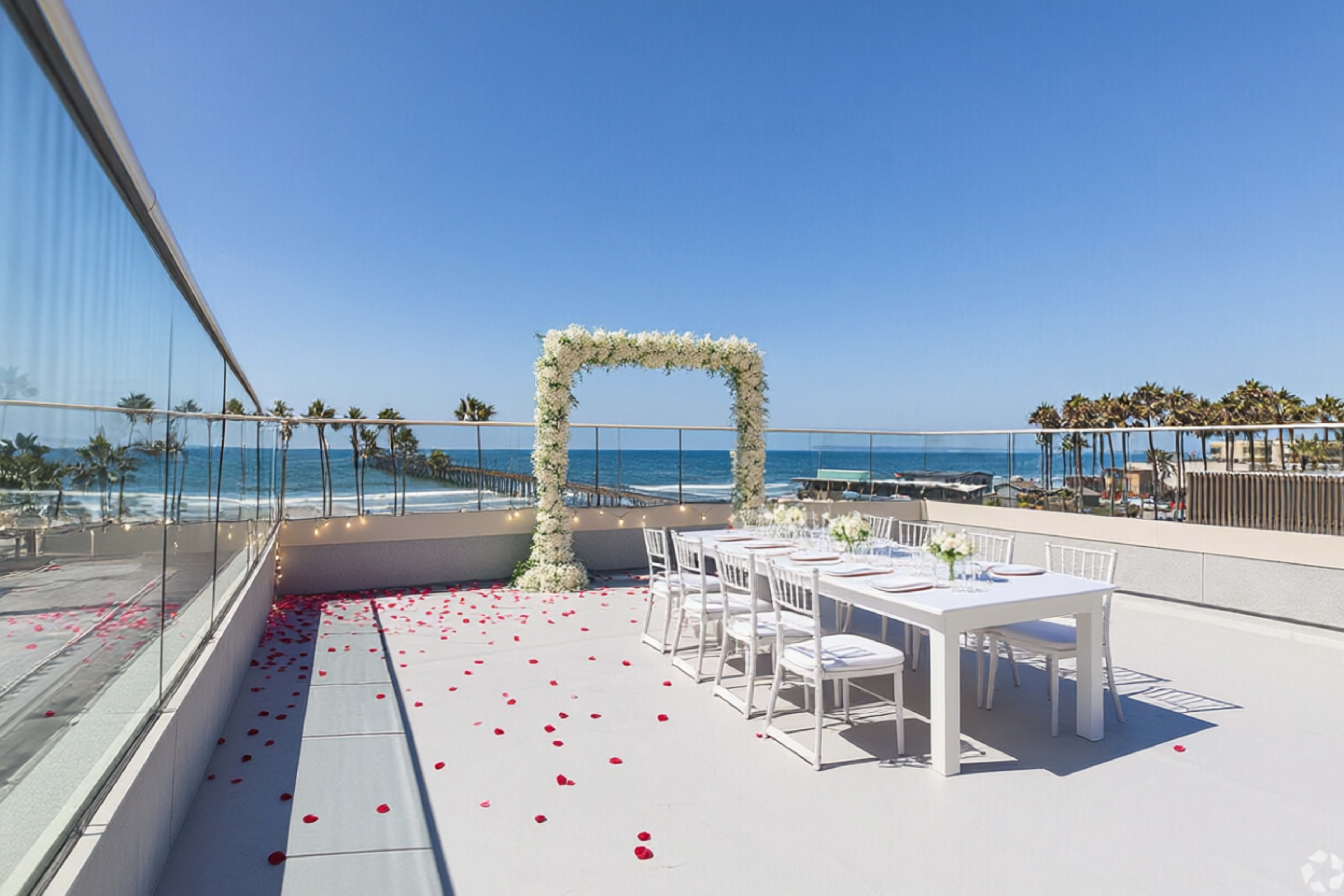 The image shows a beautifully arranged rooftop terrace overlooking the ocean, featuring a dining table set for an event under a floral arch, with rose petals scattered on the floor.