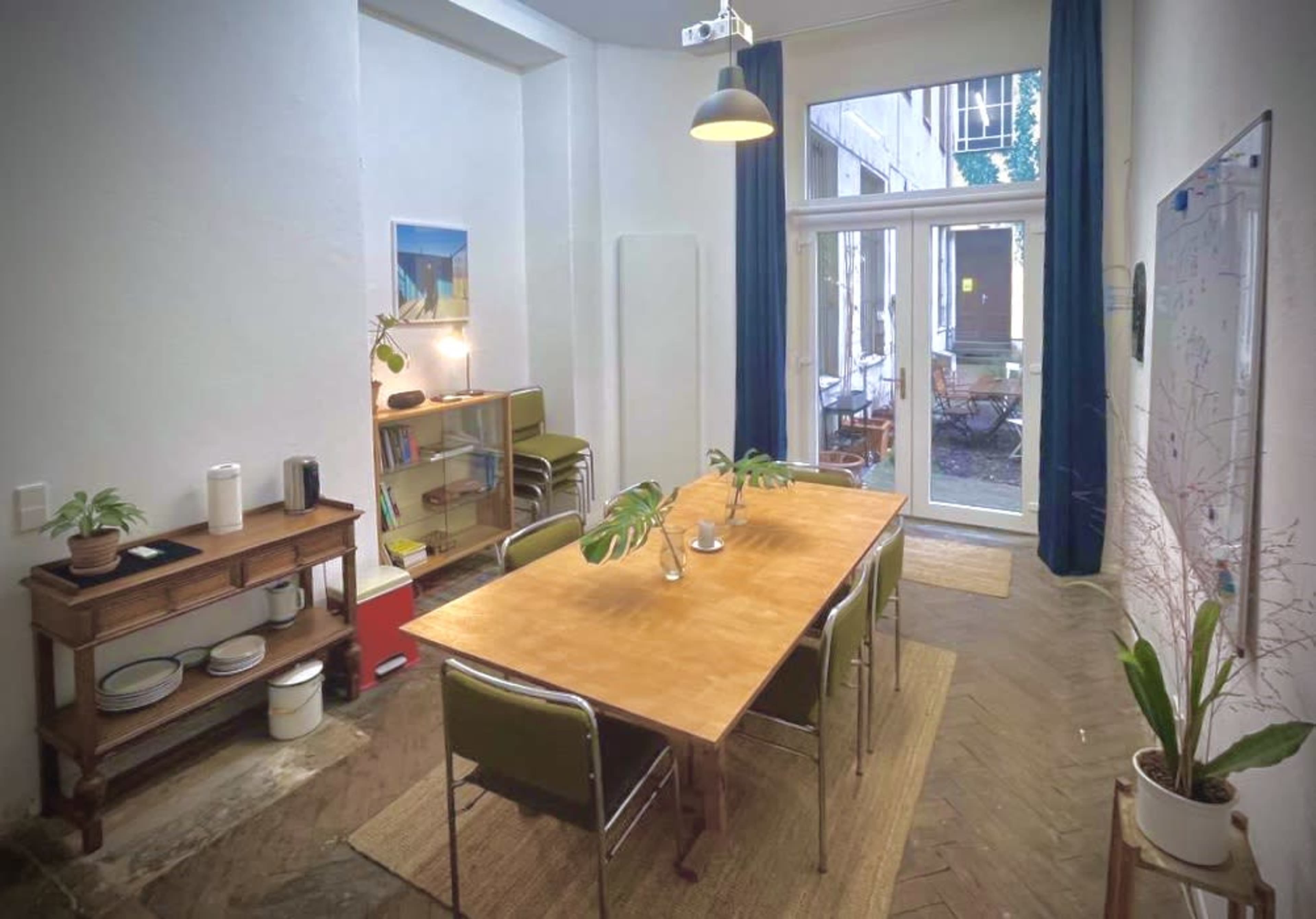 The image shows a dining area with a wooden table surrounded by chairs, a sideboard with dishes, and large windows opening to an outdoor space.