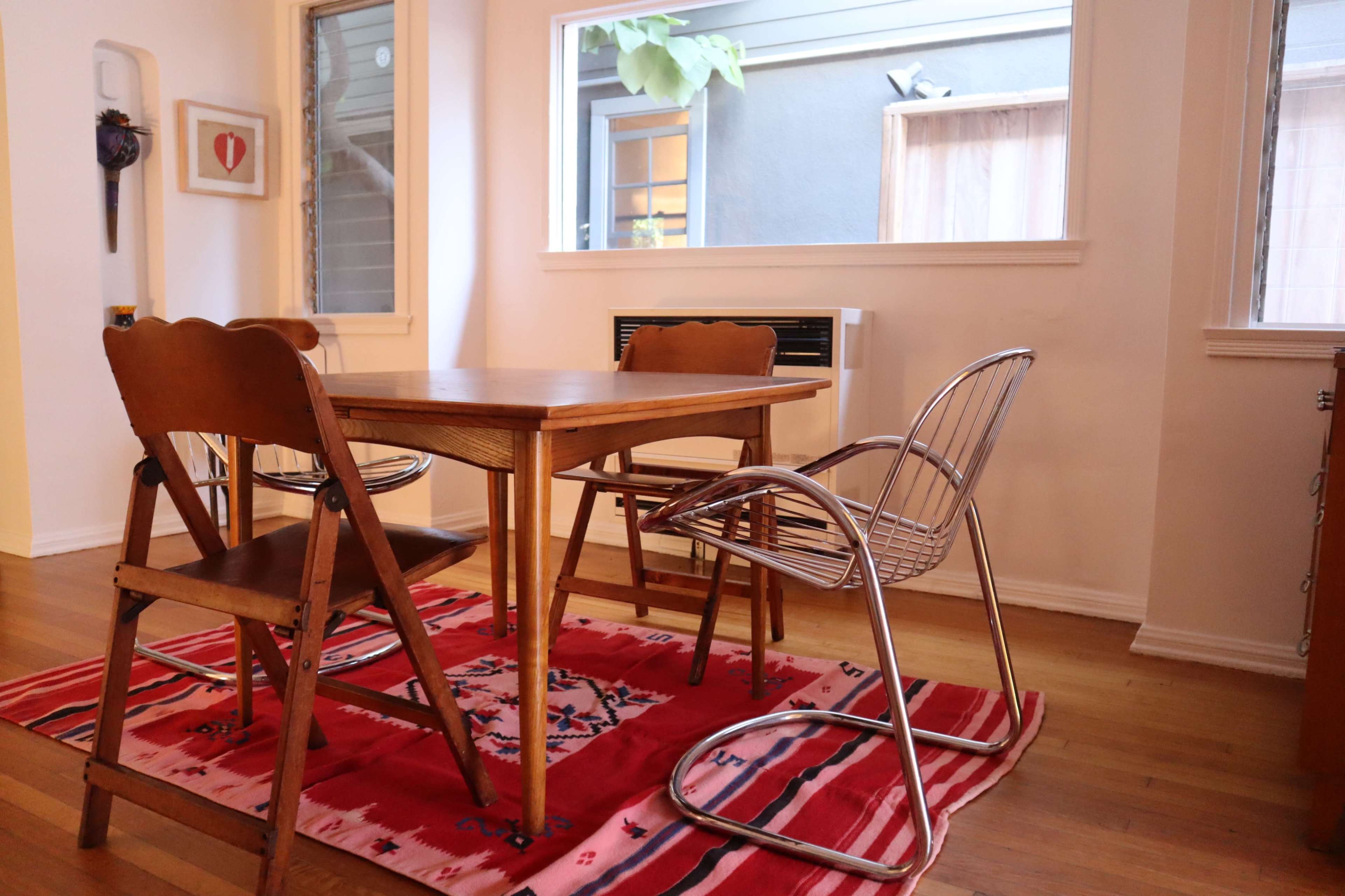 A wooden table surrounded by four chairs sits on a red rug in a well-lit dining area with a view of an outdoor space.