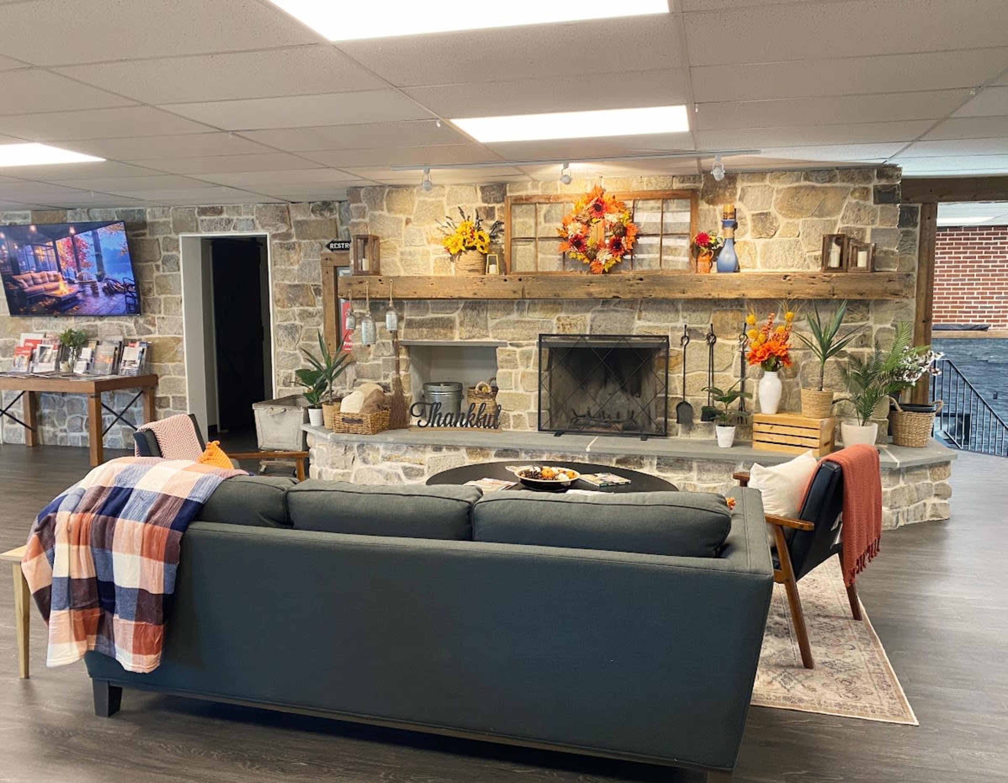 A cozy living area with a stone fireplace, a dark sofa, a patterned blanket draped over the back, and decorative plants and flowers arranged on the mantel.