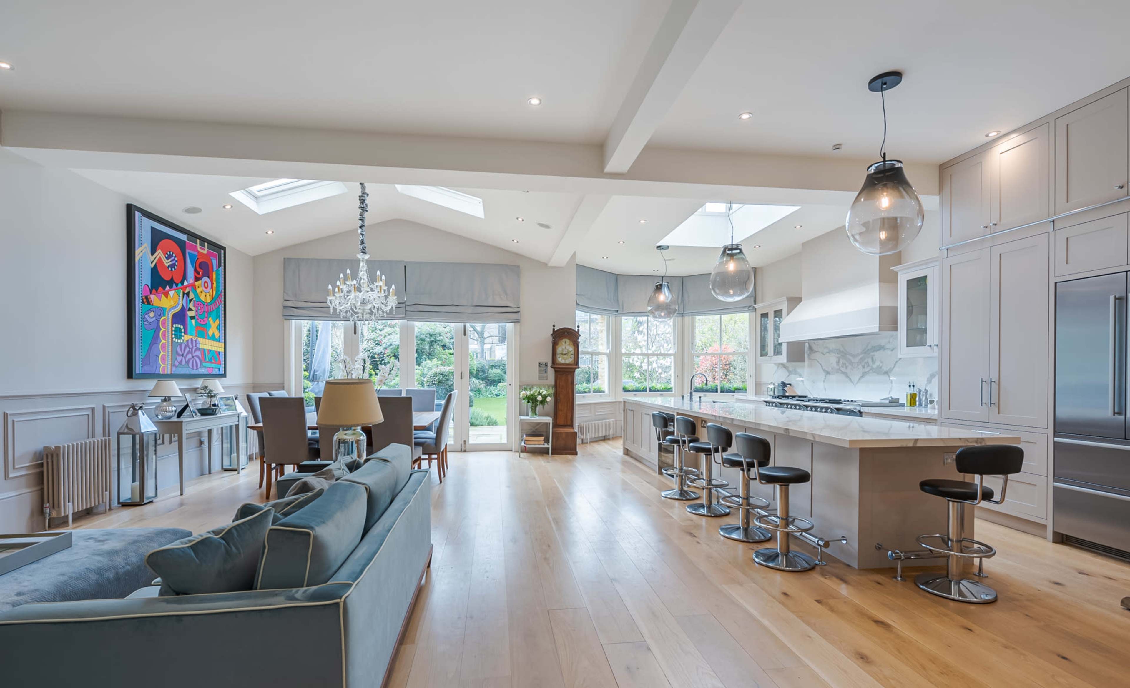 The image shows a modern open-concept kitchen and living area with large windows, a central island with bar seating, and a view of a garden.