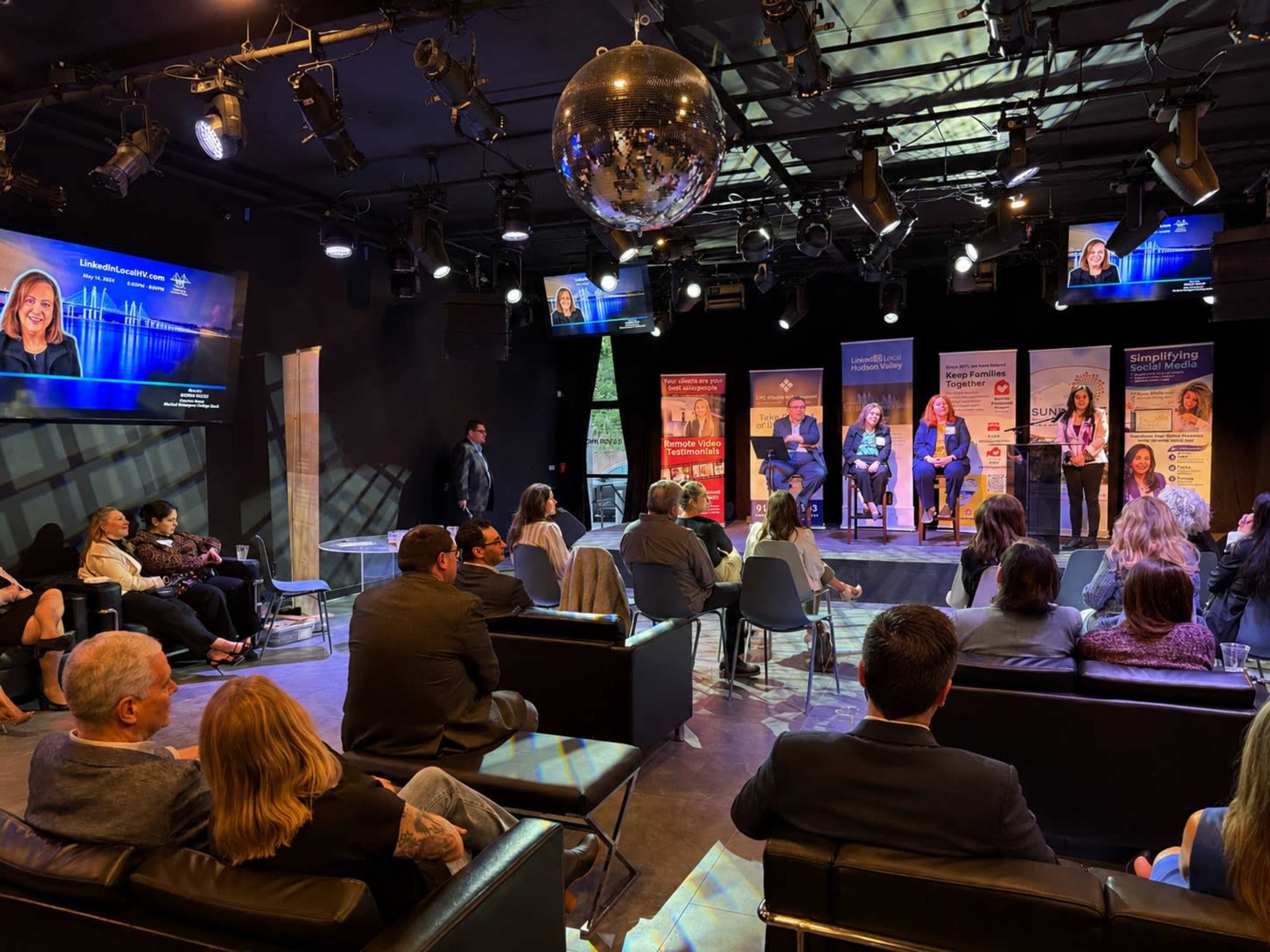 A panel discussion is taking place on a dimly lit stage with an audience seated on couches and chairs.