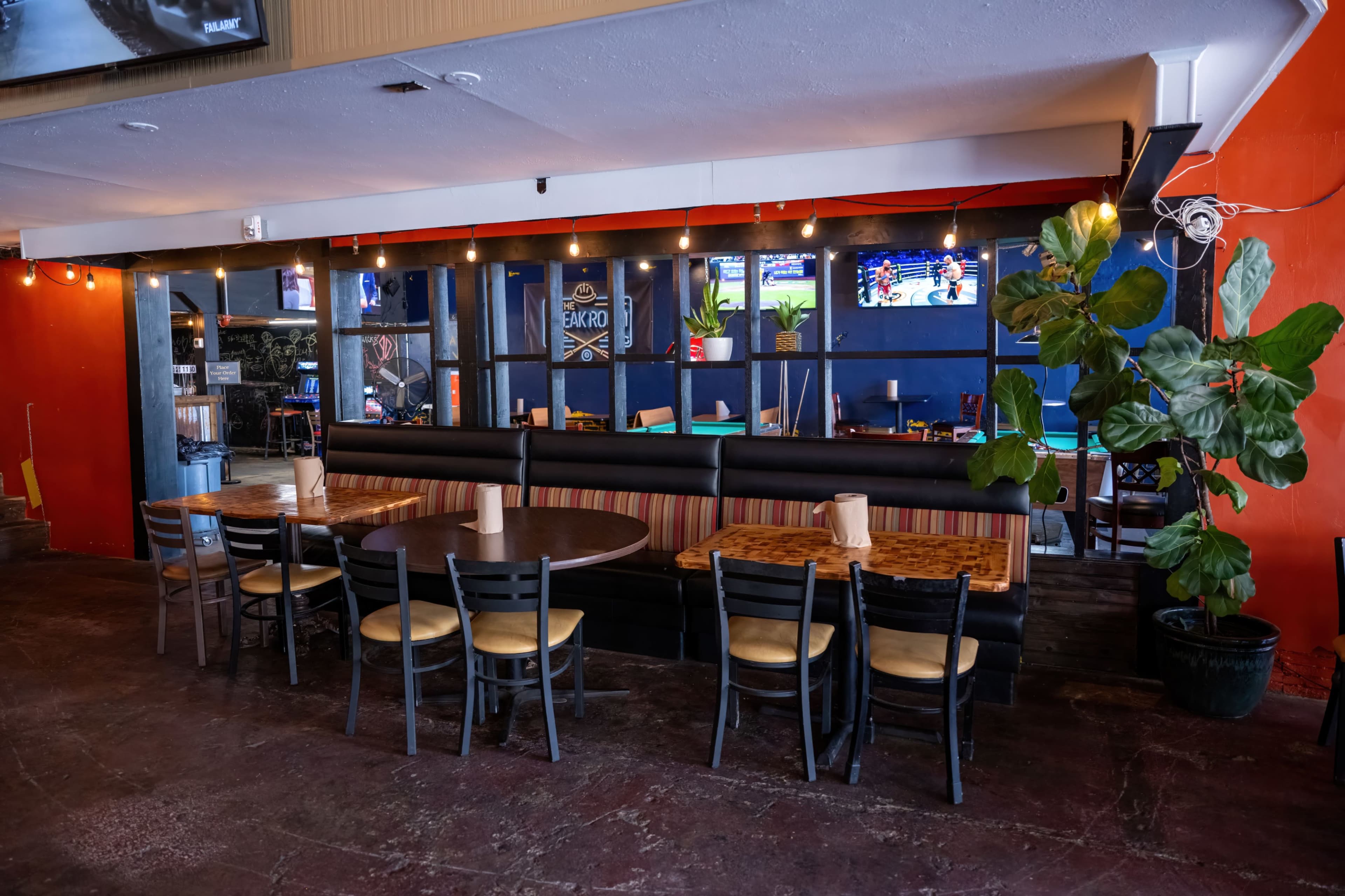 The image shows an interior view of a casual dining restaurant featuring a mix of round and rectangular tables, colorful walls, and TVs mounted on the walls.