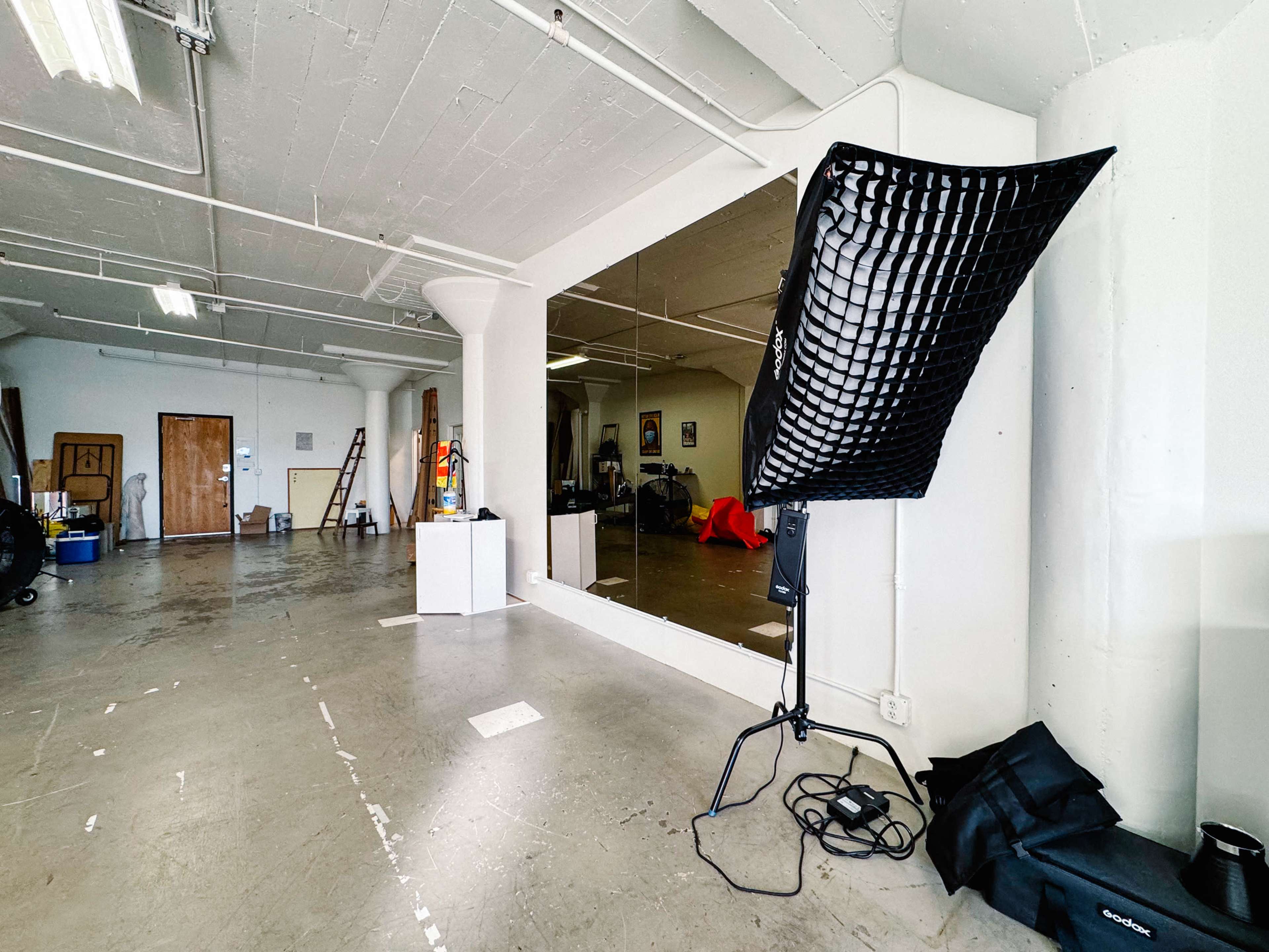 The image shows a spacious, unfinished studio with a large mirror, lighting equipment, and scattered furniture.
