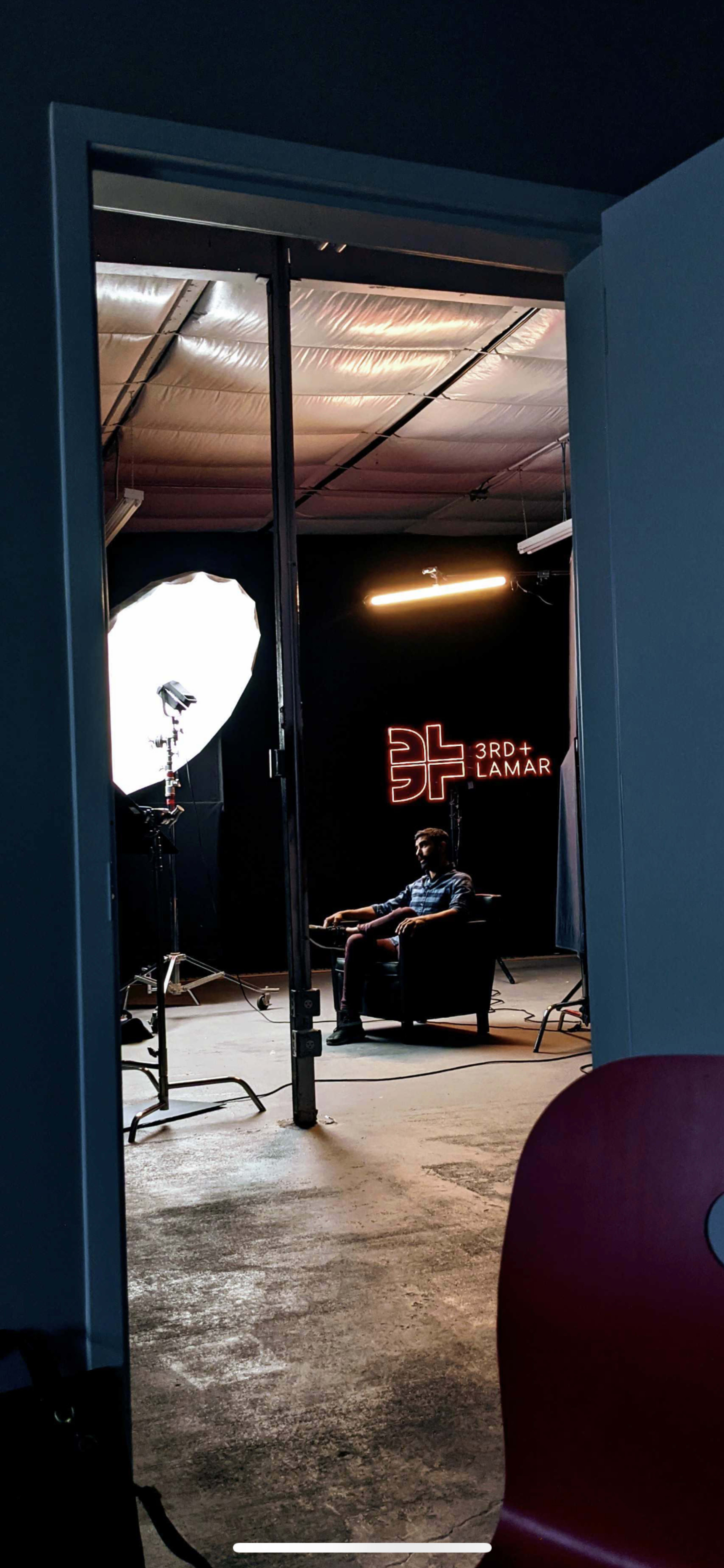The image shows a person seated in an armchair in a dimly lit studio, with a large softbox and neon signage visible in the background.