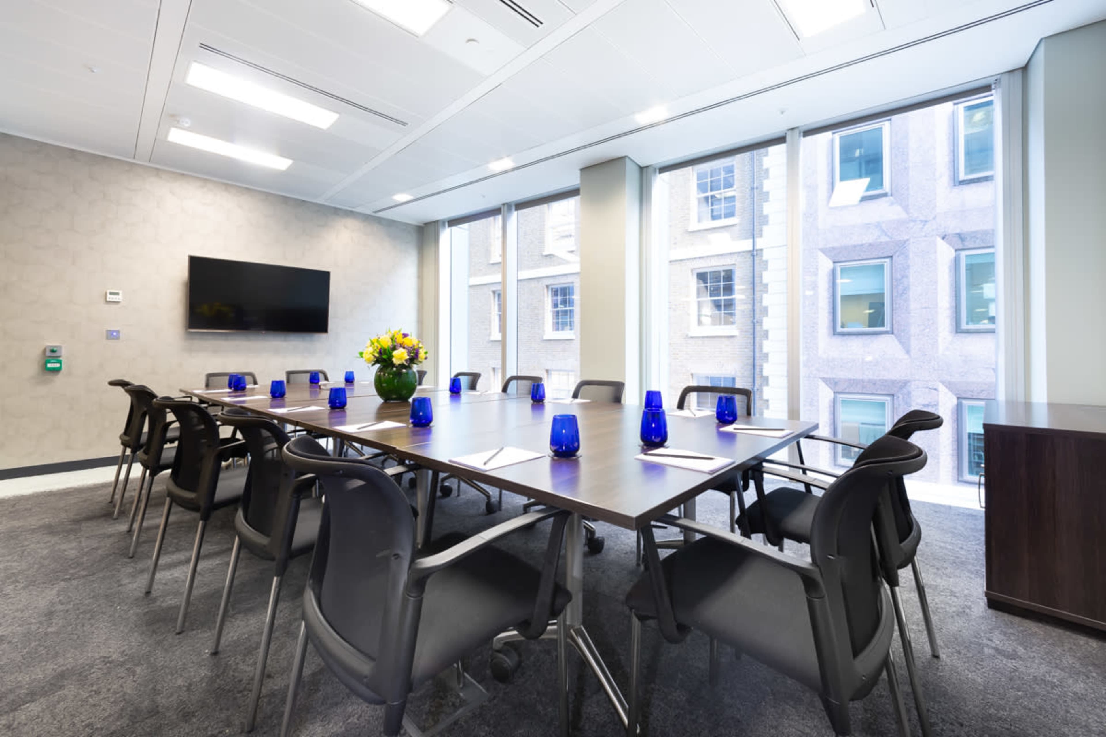 A modern conference room features a long table with blue glasses and a flower arrangement, surrounded by chairs, with large windows overlooking a building exterior.