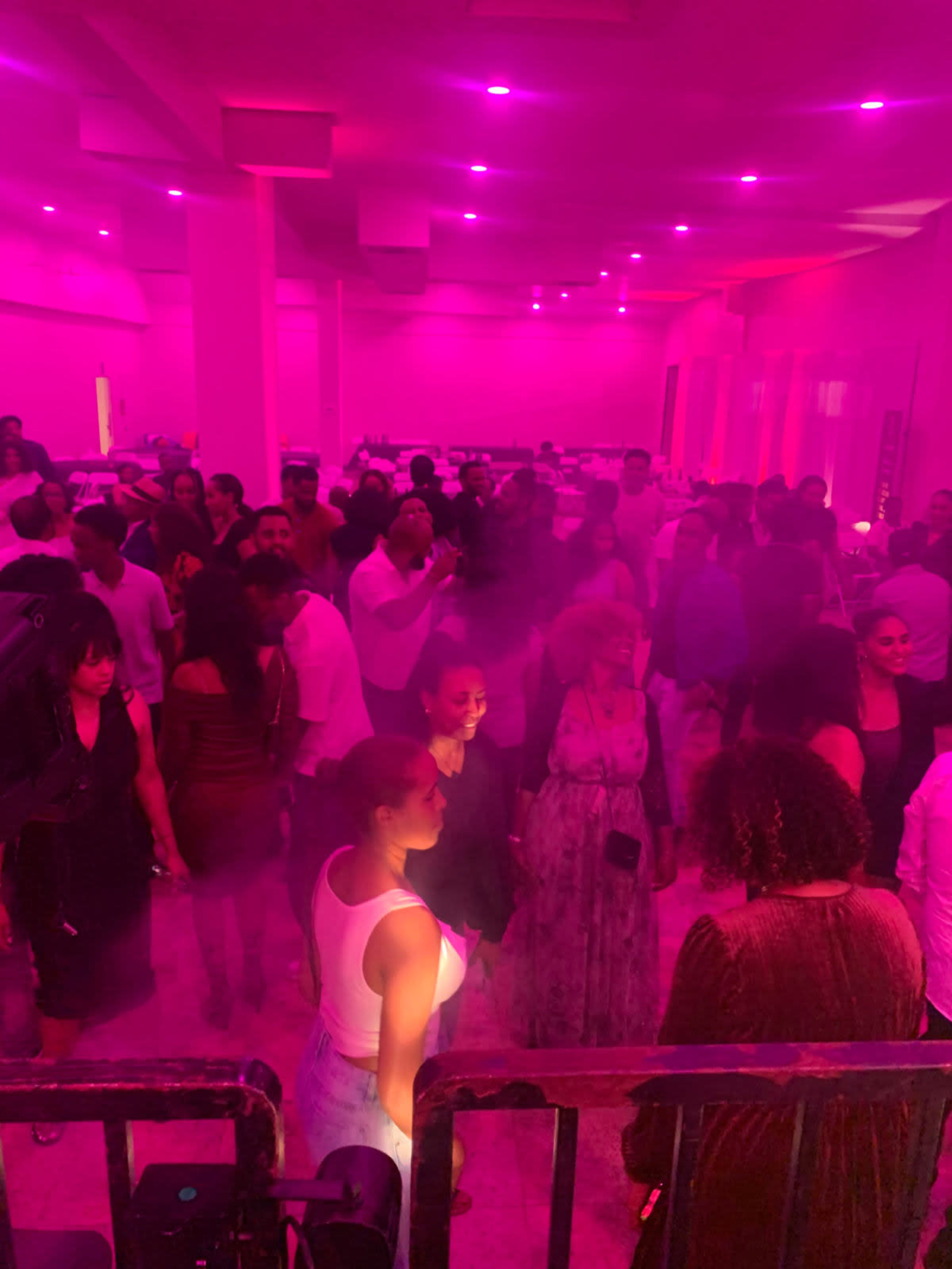 A crowded room filled with people, illuminated by bright pink lighting and a hazy atmosphere.