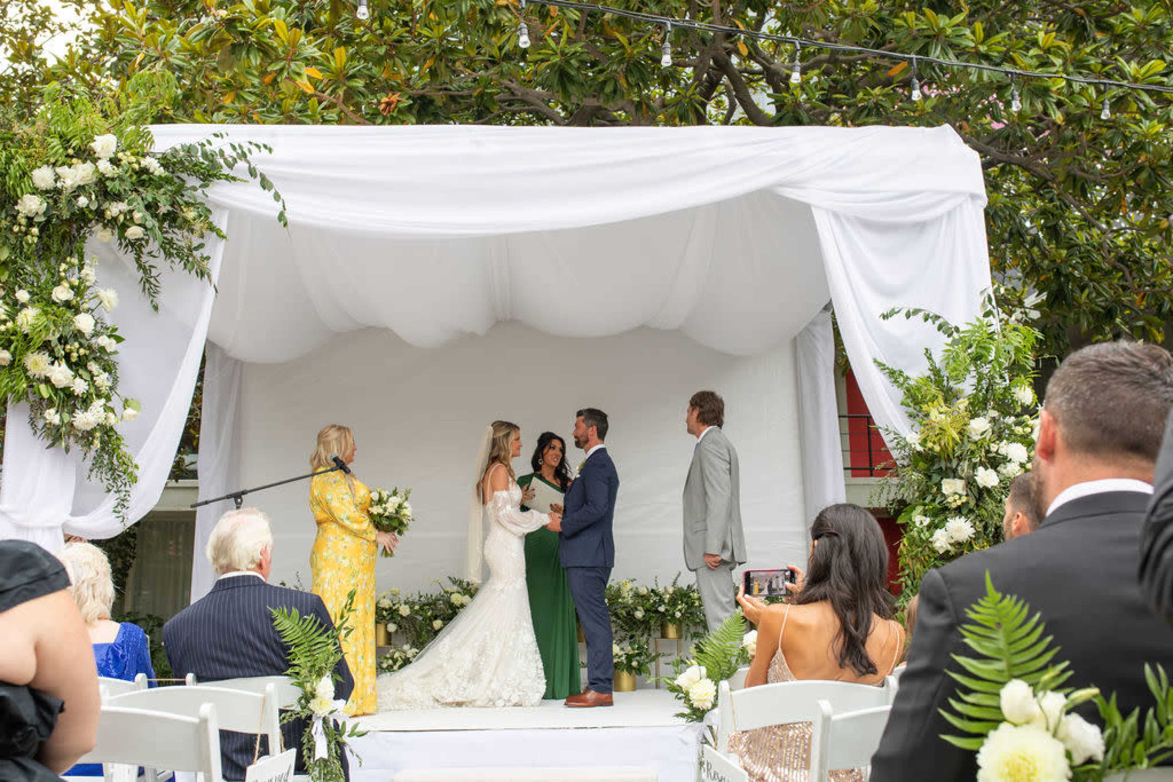 A wedding ceremony is taking place outdoors under a white draped arch, with the couple exchanging vows in front of guests.