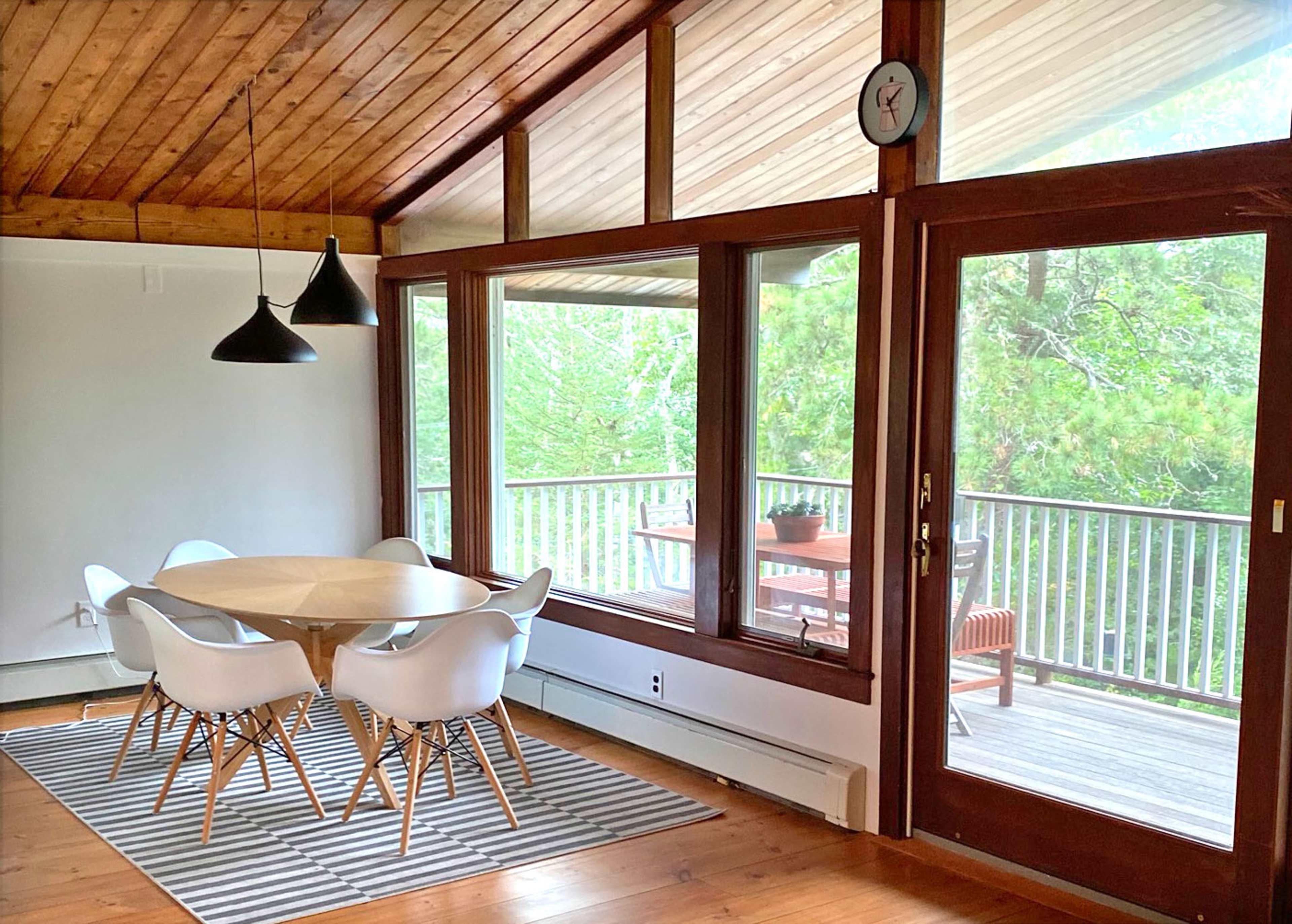 A dining area features a circular wooden table surrounded by white chairs, with large windows overlooking a deck and trees.