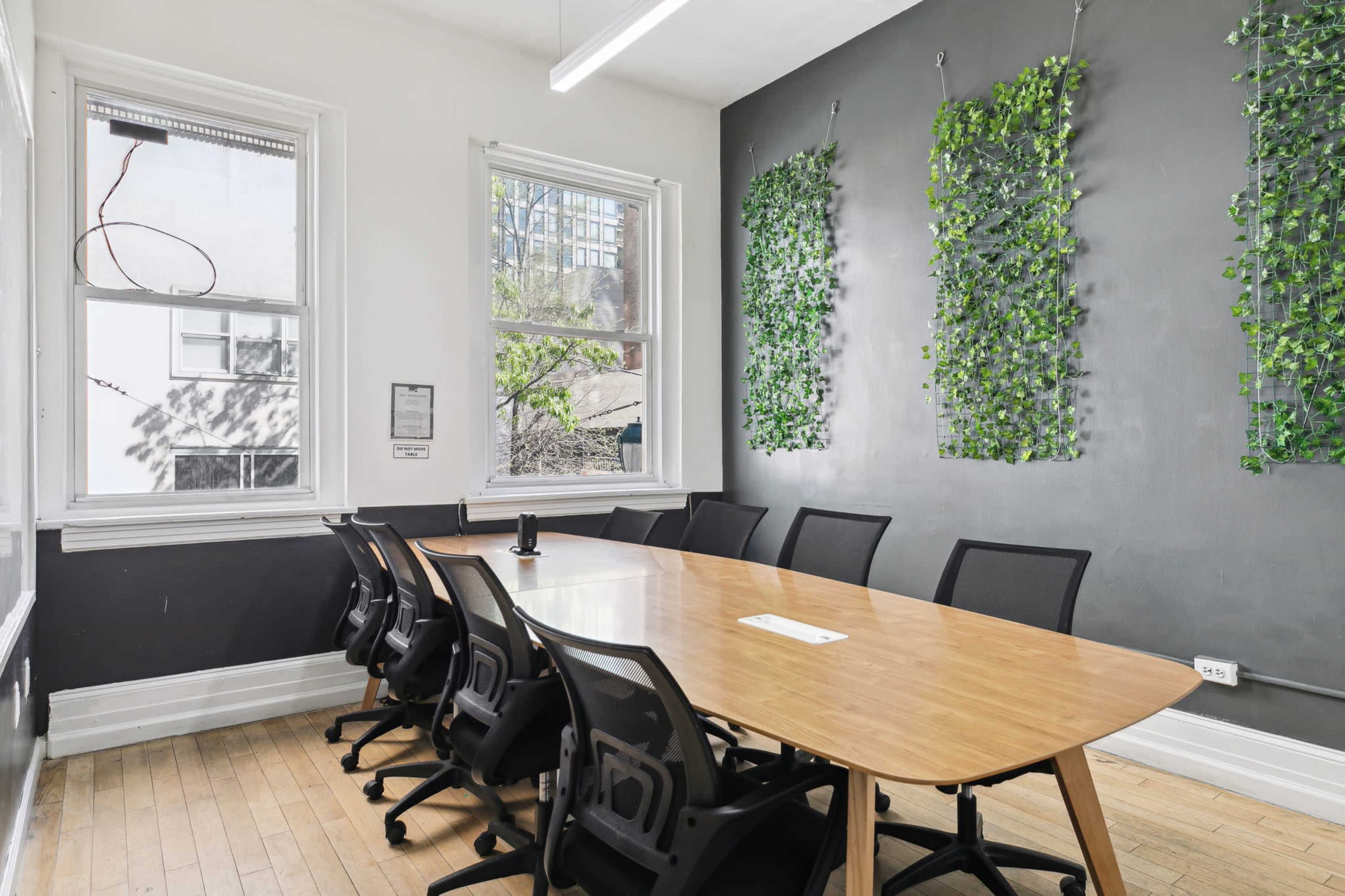 A conference room features a long wooden table surrounded by black chairs, with green wall decor and large windows.