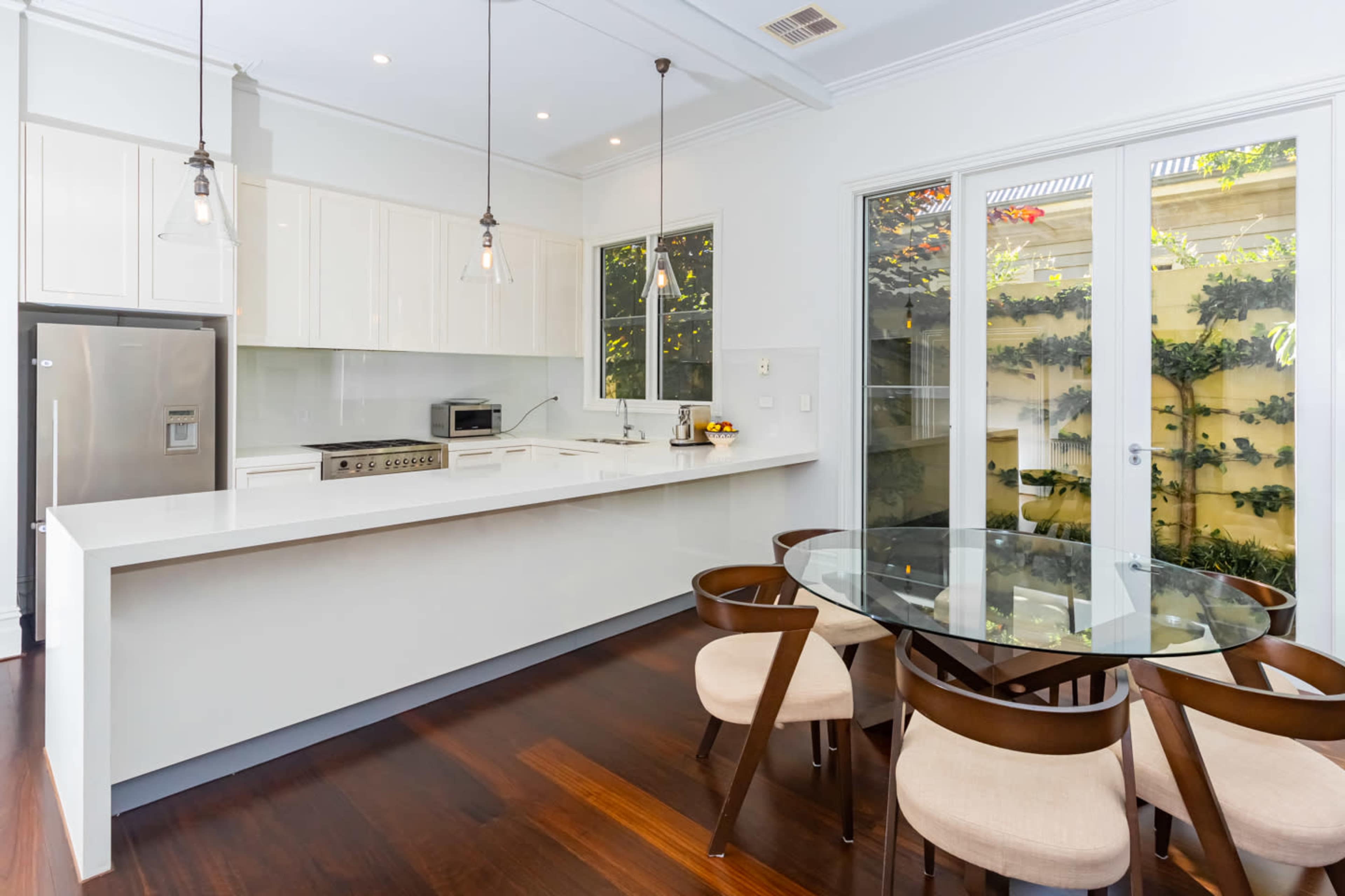 The image shows a modern kitchen area with white cabinetry, stainless steel appliances, and a glass dining table surrounded by wooden chairs.