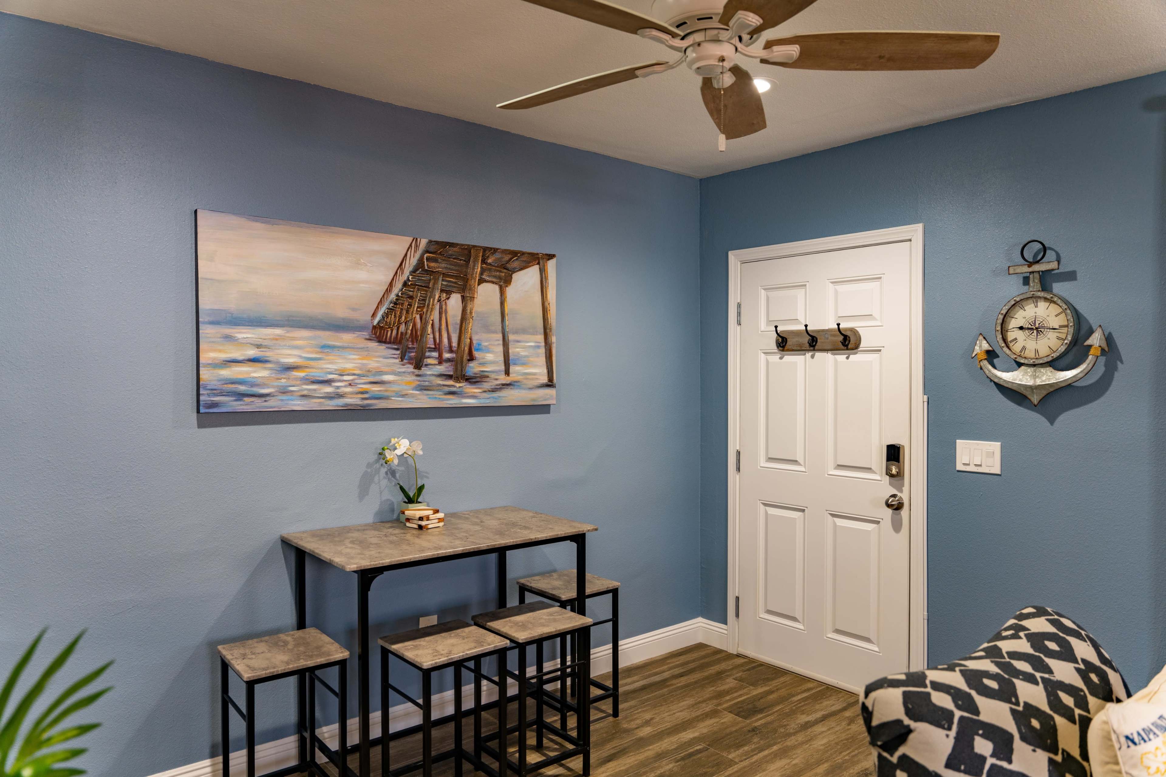 The image shows a small dining area with a table and four stools against a blue wall, complemented by a painting of a pier and a clock on the adjacent wall.