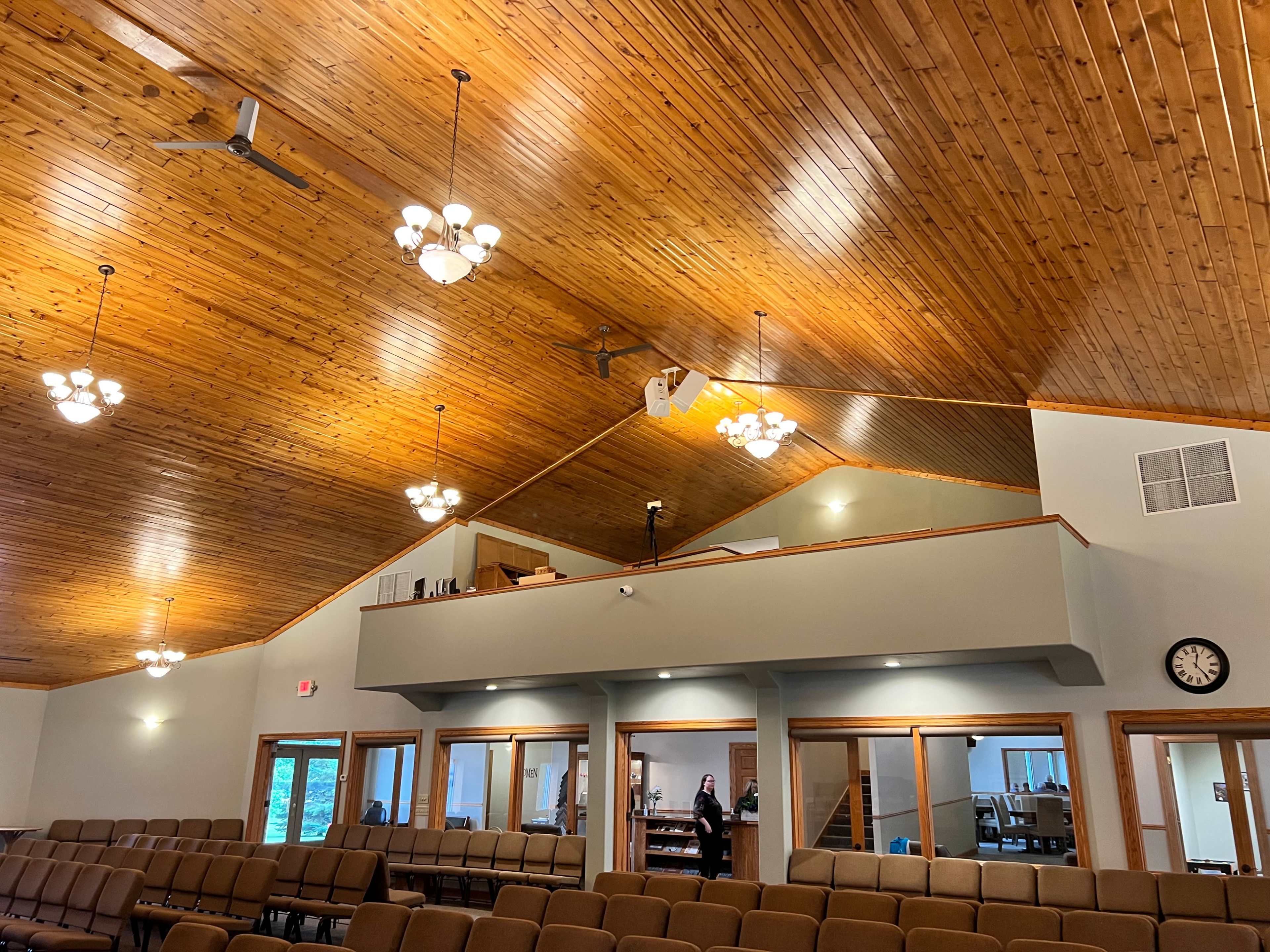 The interior of a church with wooden beams, overhead lights, and rows of chairs facing a stage area.