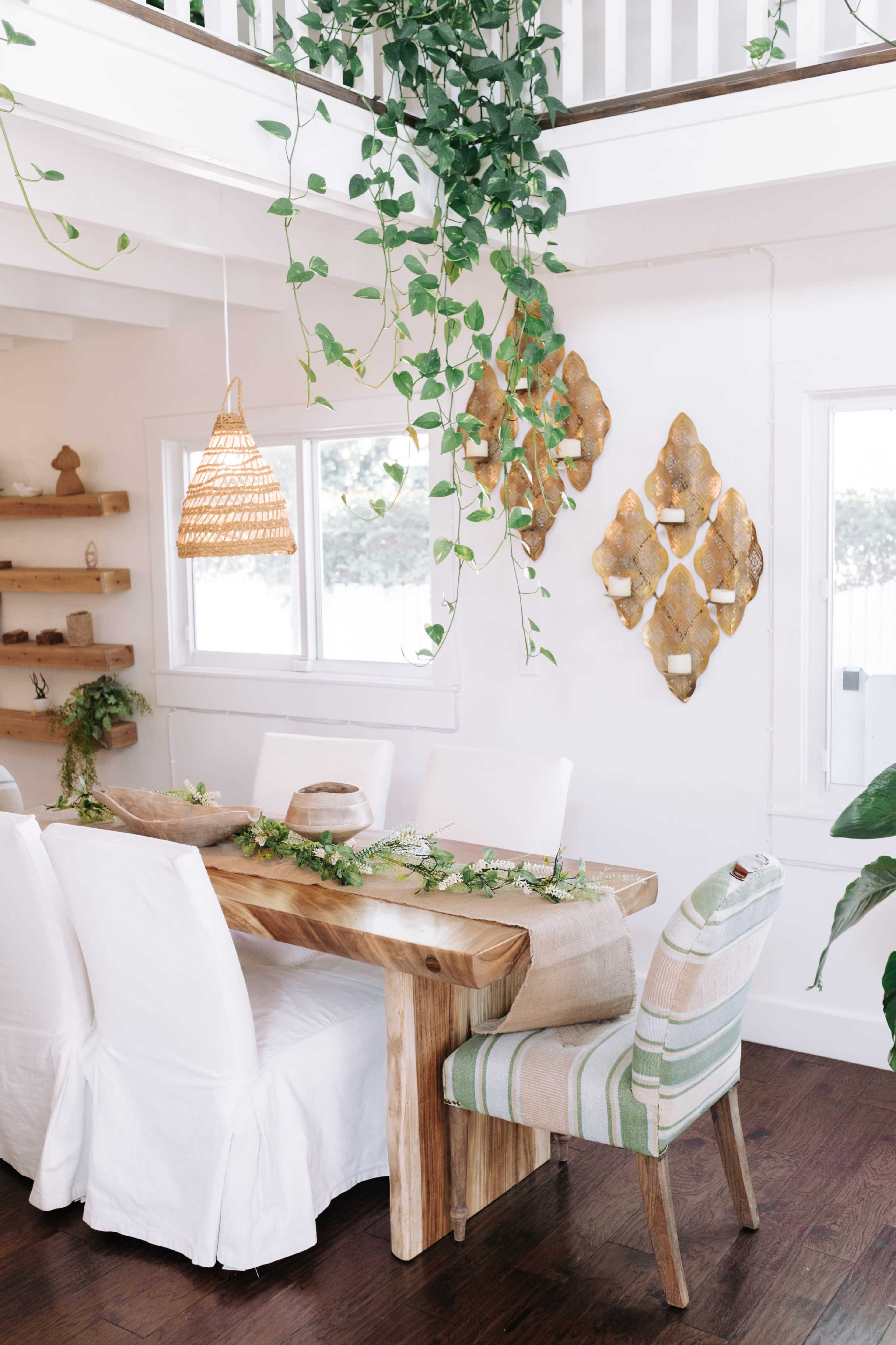 A wooden dining table is set in a well-lit room with white walls, surrounded by white chairs and accented by hanging plants and decorative wall art.