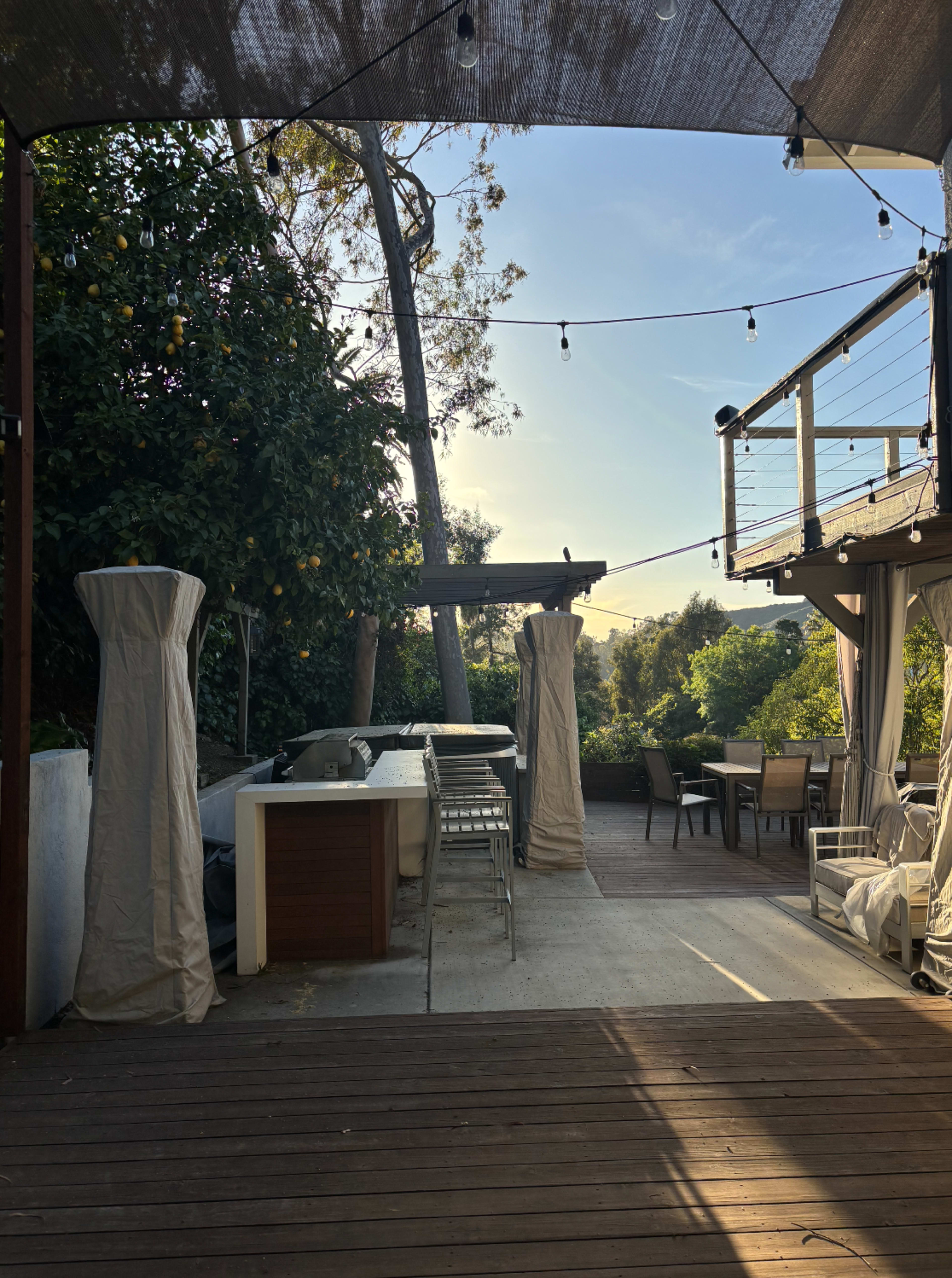 The image shows an outdoor patio area with a bar, dining table, and string lights, surrounded by greenery and covered by a canopy.