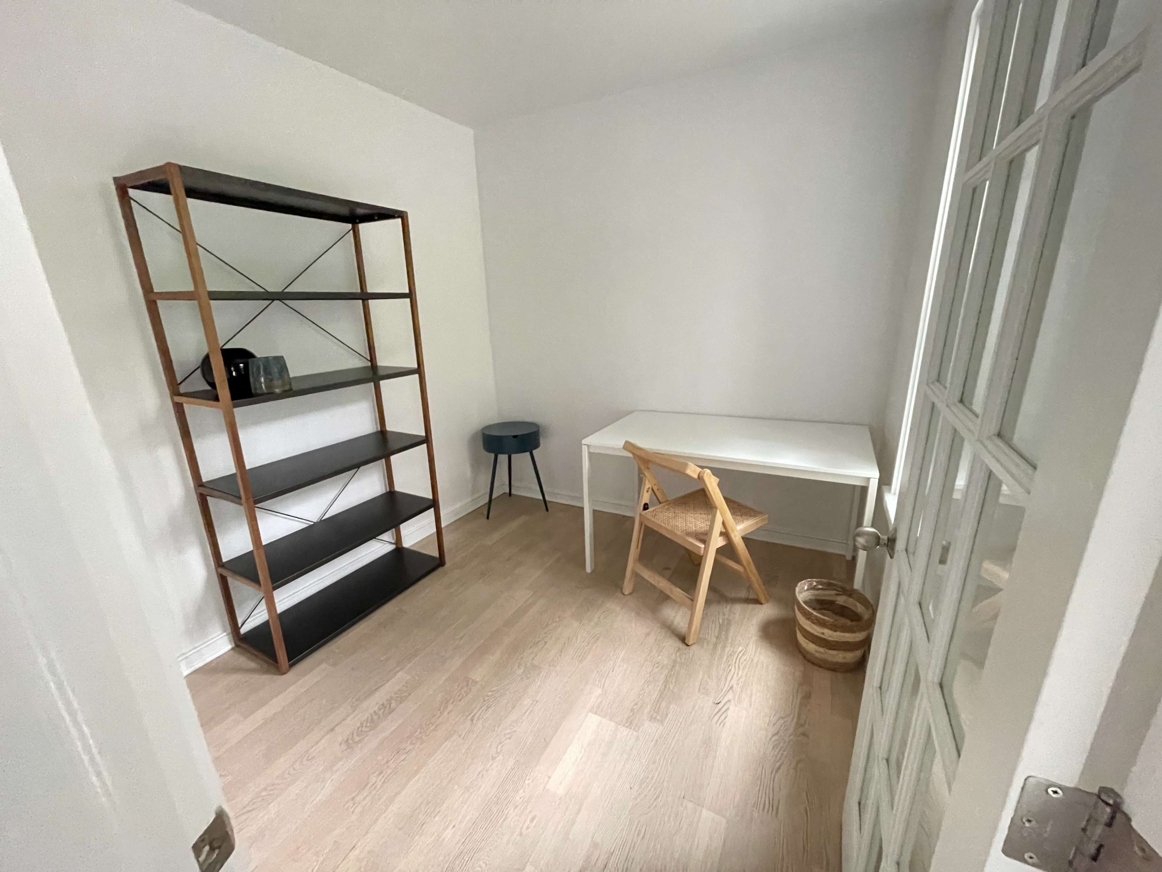 The image shows a minimalist room featuring a tall bookshelf, a small blue stool, a white desk, and a woven basket on the floor.