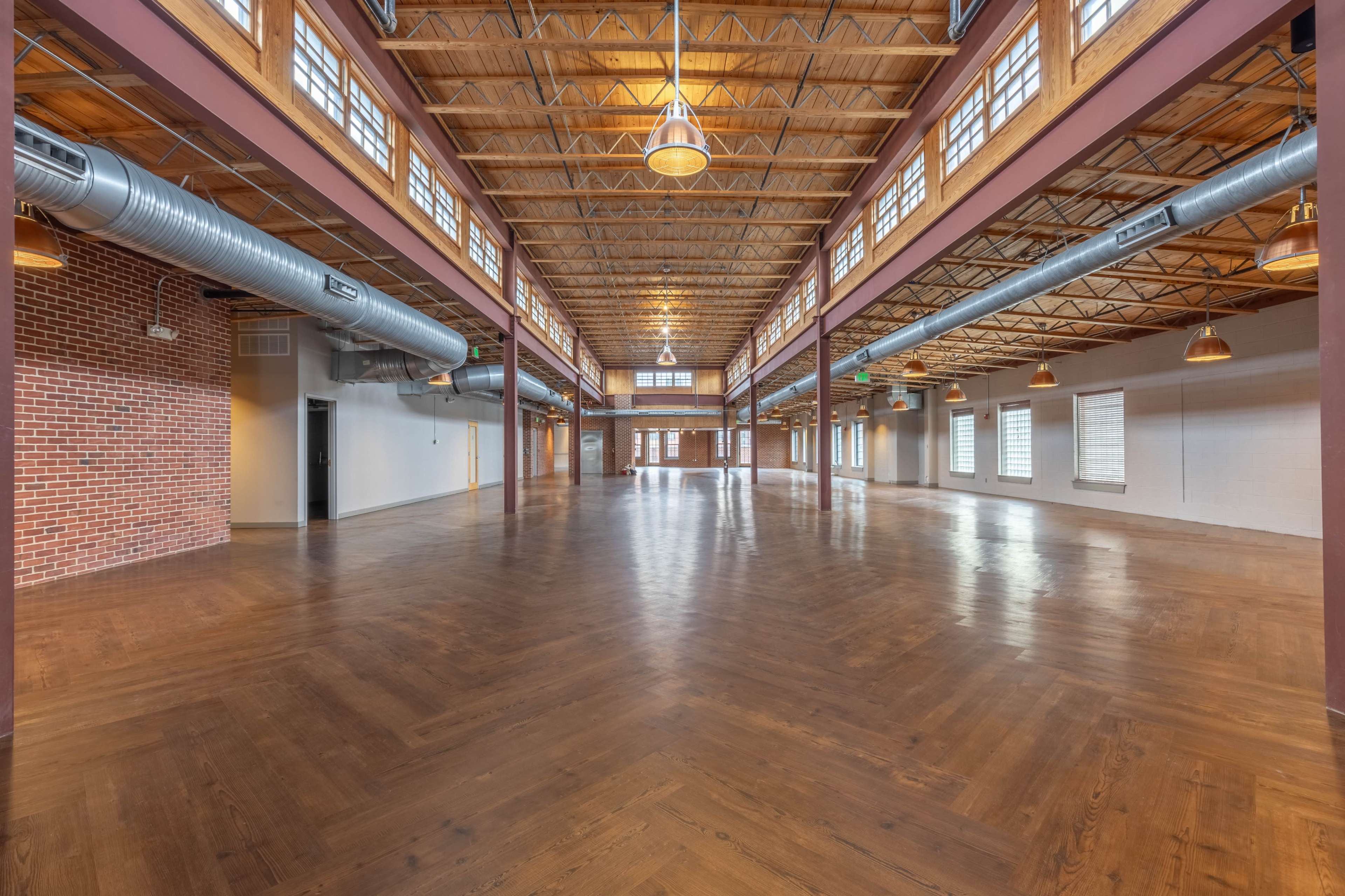 The image shows a spacious, open indoor venue with wooden floors, exposed wooden beams, and large windows along the walls.