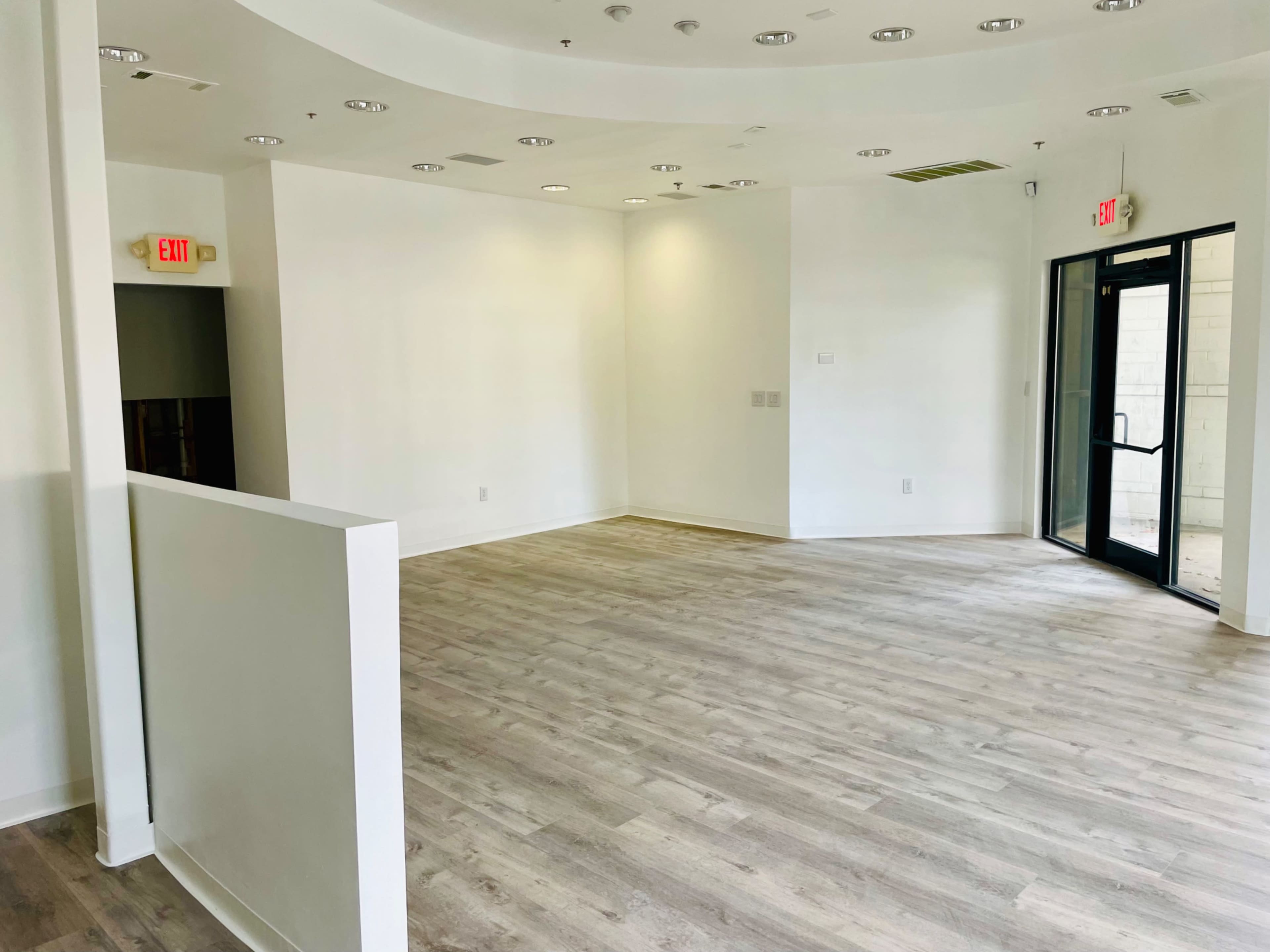 An empty, well-lit room with light wooden flooring, white walls, and a glass entrance.